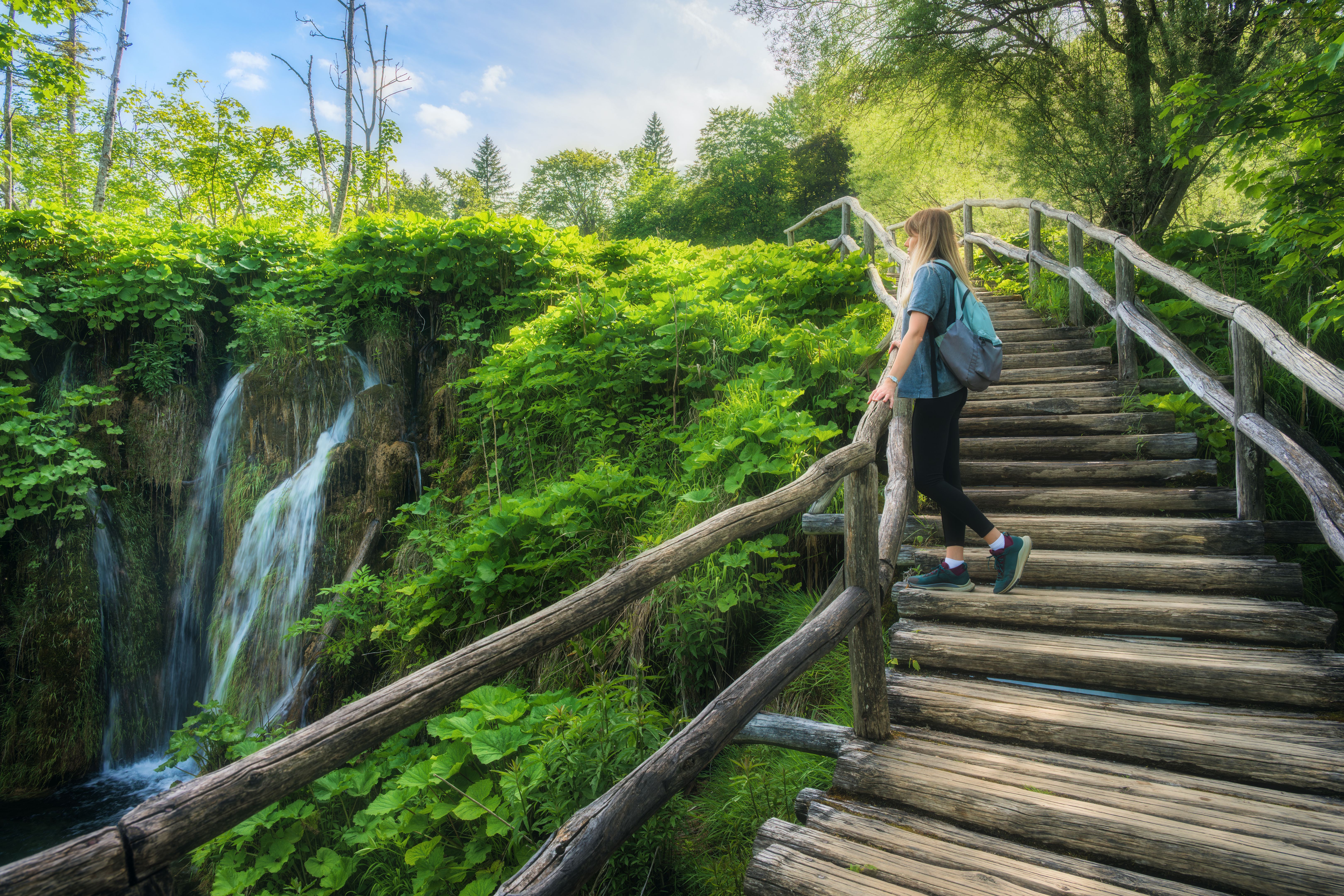 Young woman with backpack walking along a wooden path near stunning waterfalls, green trees in Plitvice Lakes national park, Croatia at sunset in summer. Female tourist. Sporty girl in forest. Hiking