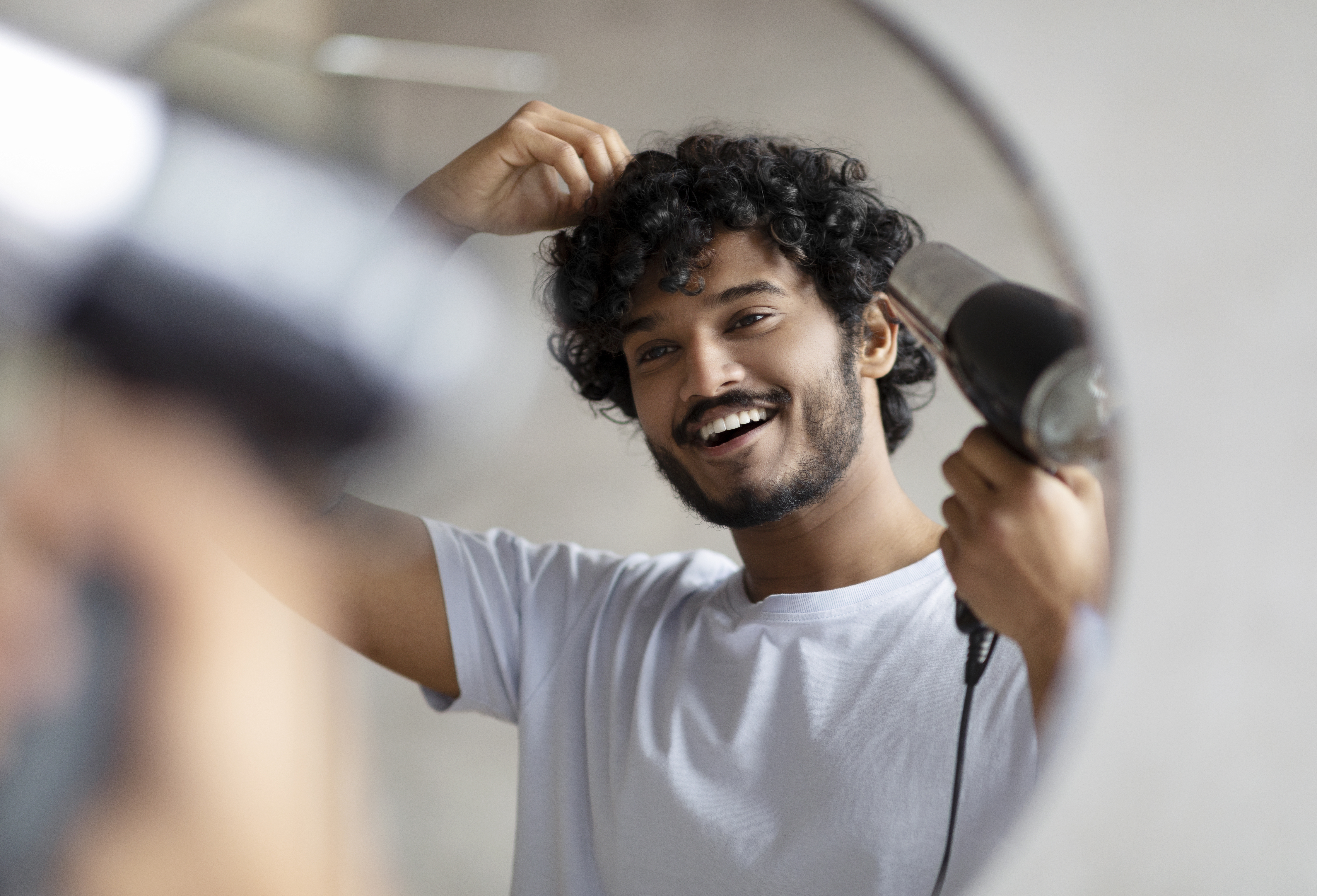 Homem indiano animado usando secador de cabelo após o banho, secando o cabelo encaracolado, fazendo penteado, fazendo rotina matinal
