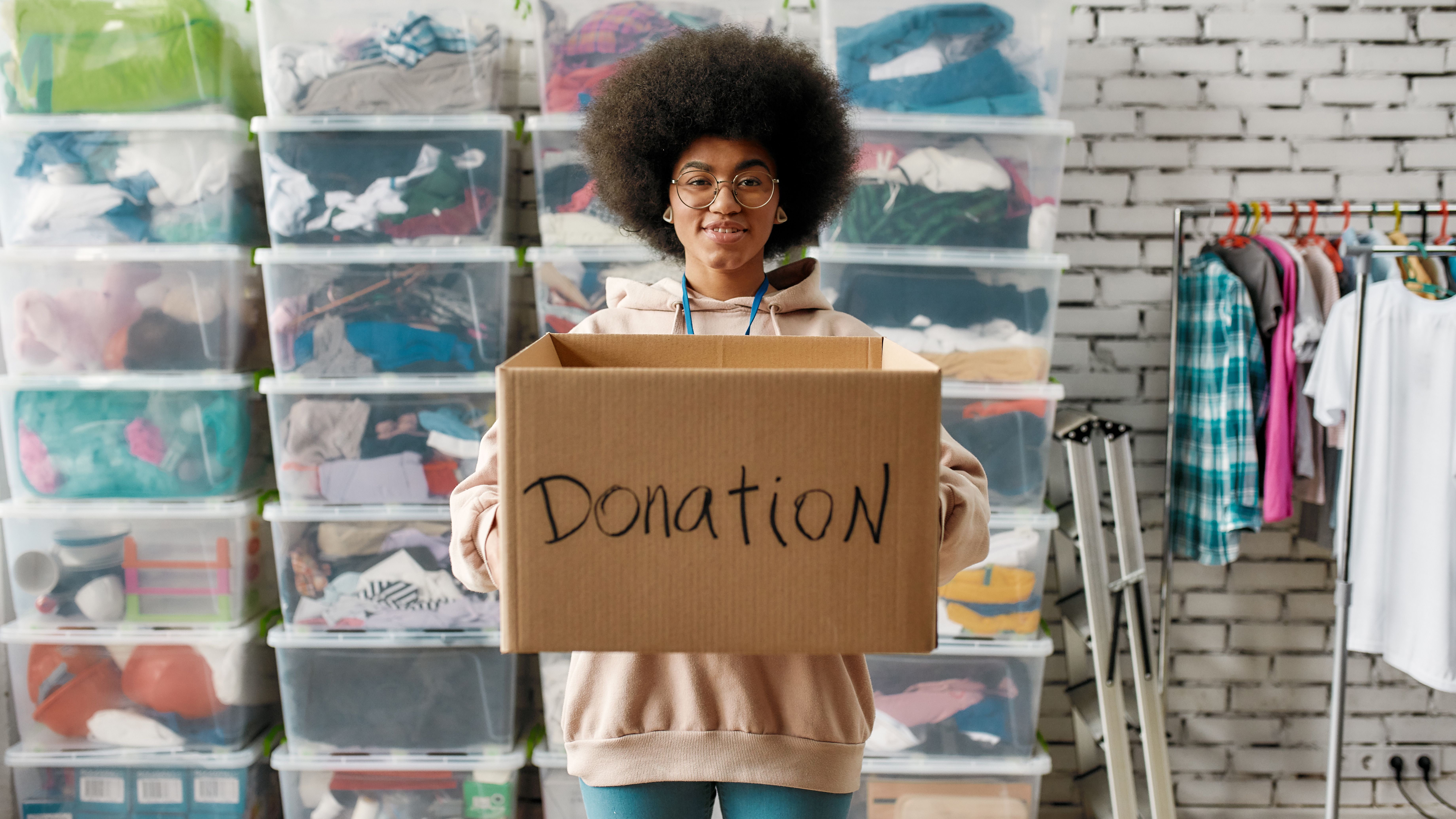 African american girl holding donation box and looking at camera, posing in front of boxes full of clothes, Young volunteer working for a charity