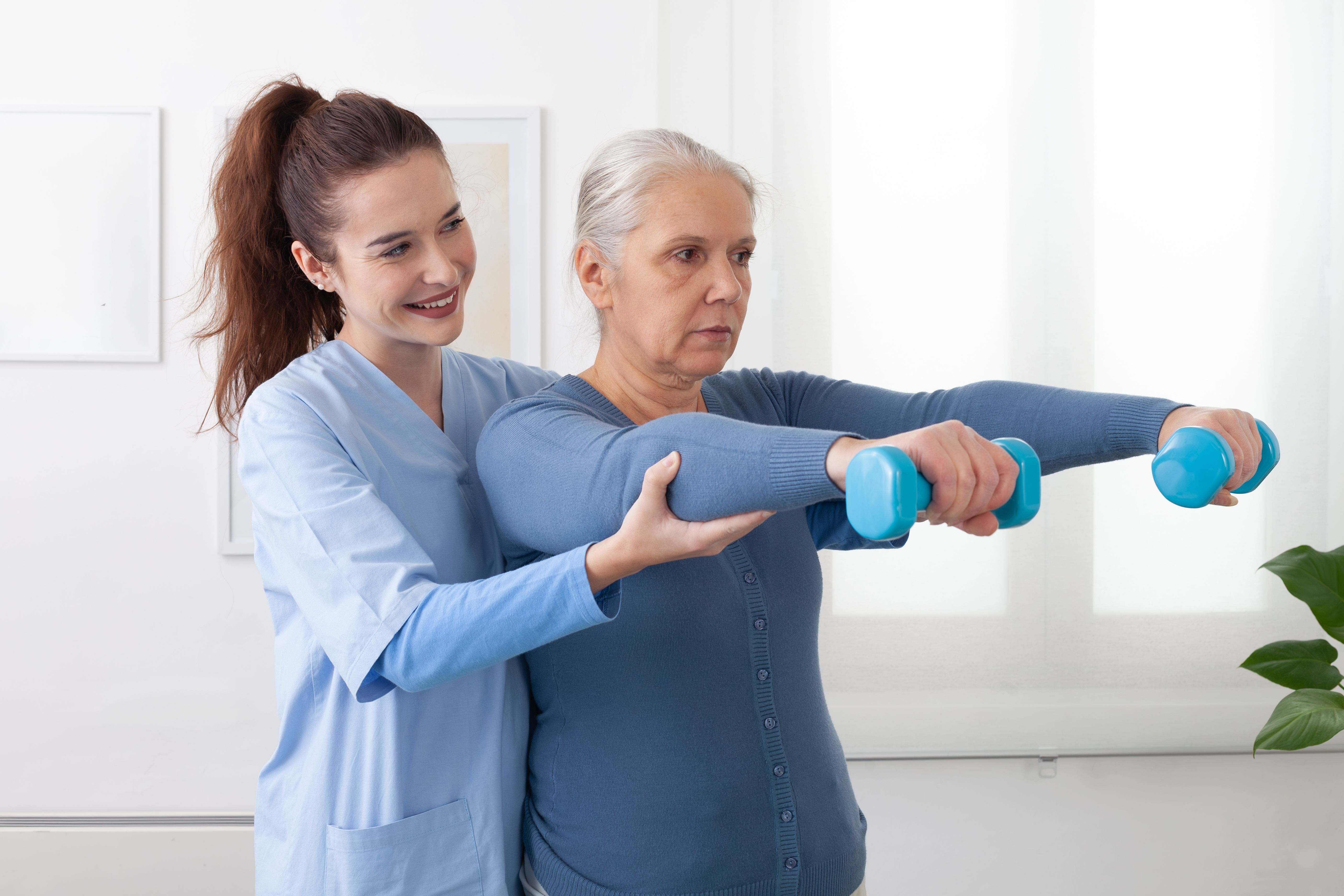 Smiling young nurse assisting an elderly woman with light dumbbell exercises during a physiotherapy session at a nursing home. The senior patient looks happy and engaged, support in a healthcare. Smiling young nurse assisting an elderly woman with light dumbbell exercises during a physiotherapy session at a nursing home. The senior patient looks happy and engaged, support in a healthcare.