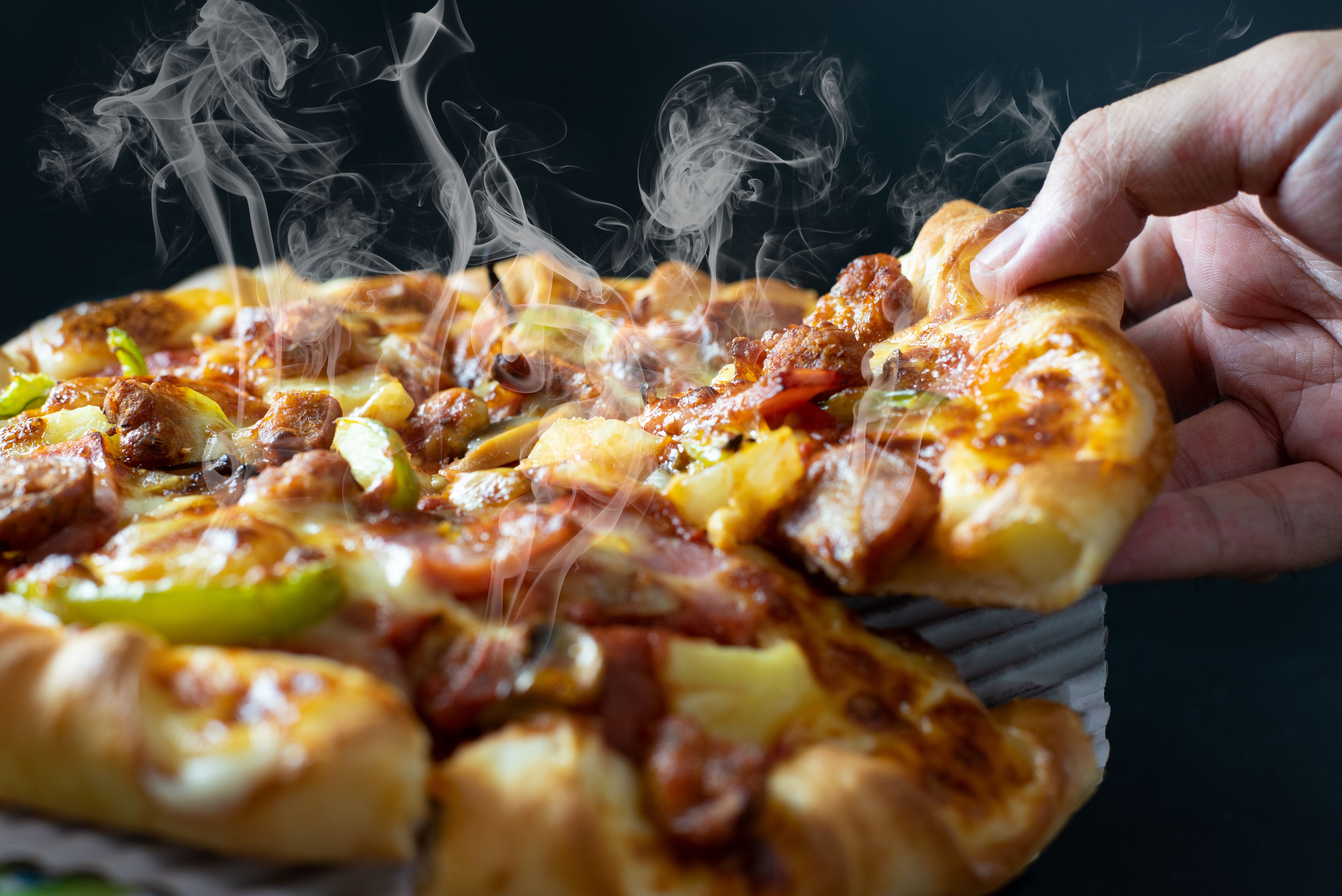 Hand picking sliced Pizza with cheese ham bacon and pepperoni on black background with hot steaming smoke. Food and cooking concept. Lunch time serve and hungry theme. Pizza delivery service to home.
