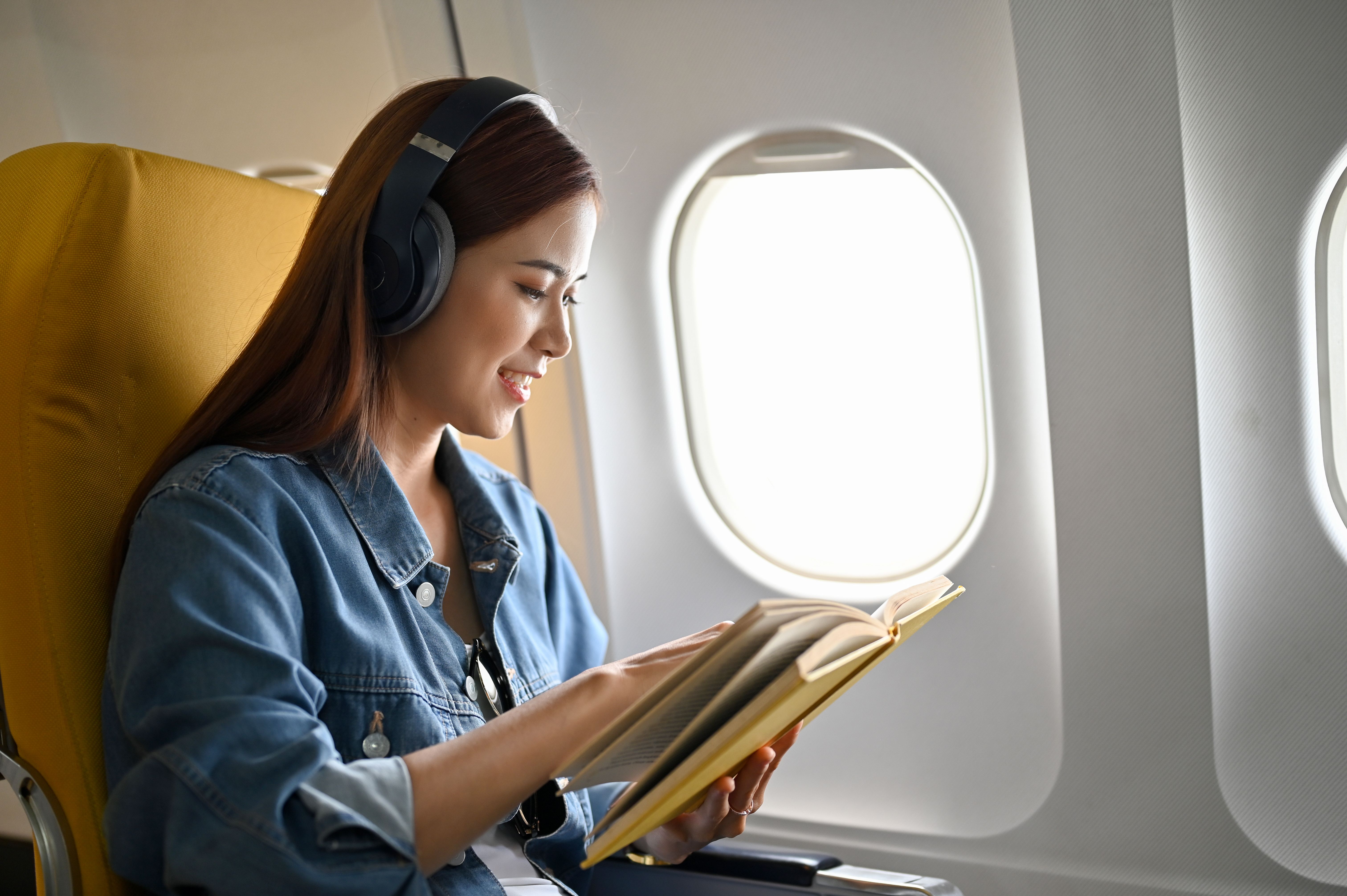 Relaxed millennial Asian female traveler with headphones reading a book on the plane Relaxed millennial Asian female traveler with headphones reading a book on the plane
