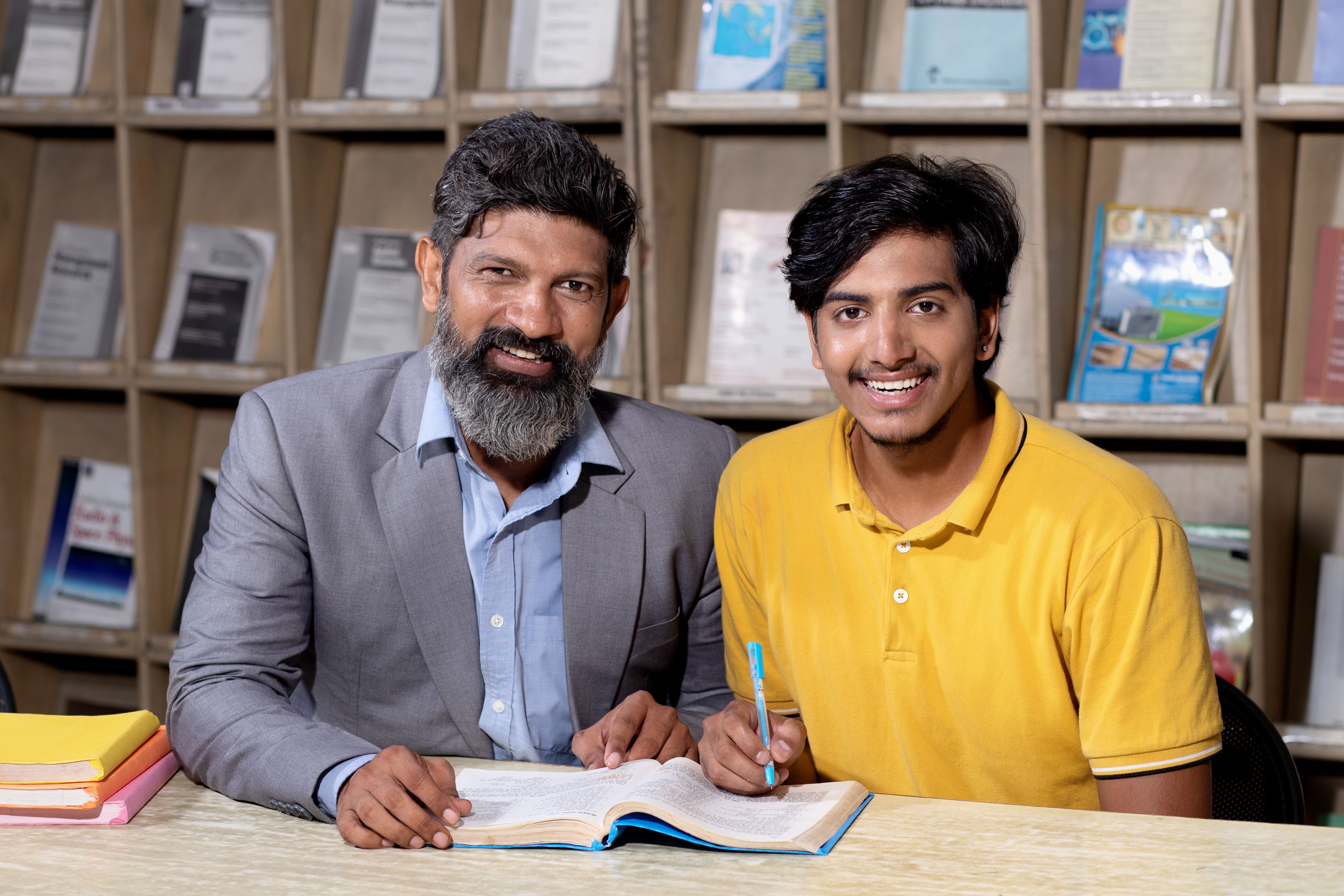 Young indian student boy and mature beard teacher reading book studying in college library with bookshelf behind. Young indian student boy and mature beard teacher reading book studying in college library with bookshelf behind.