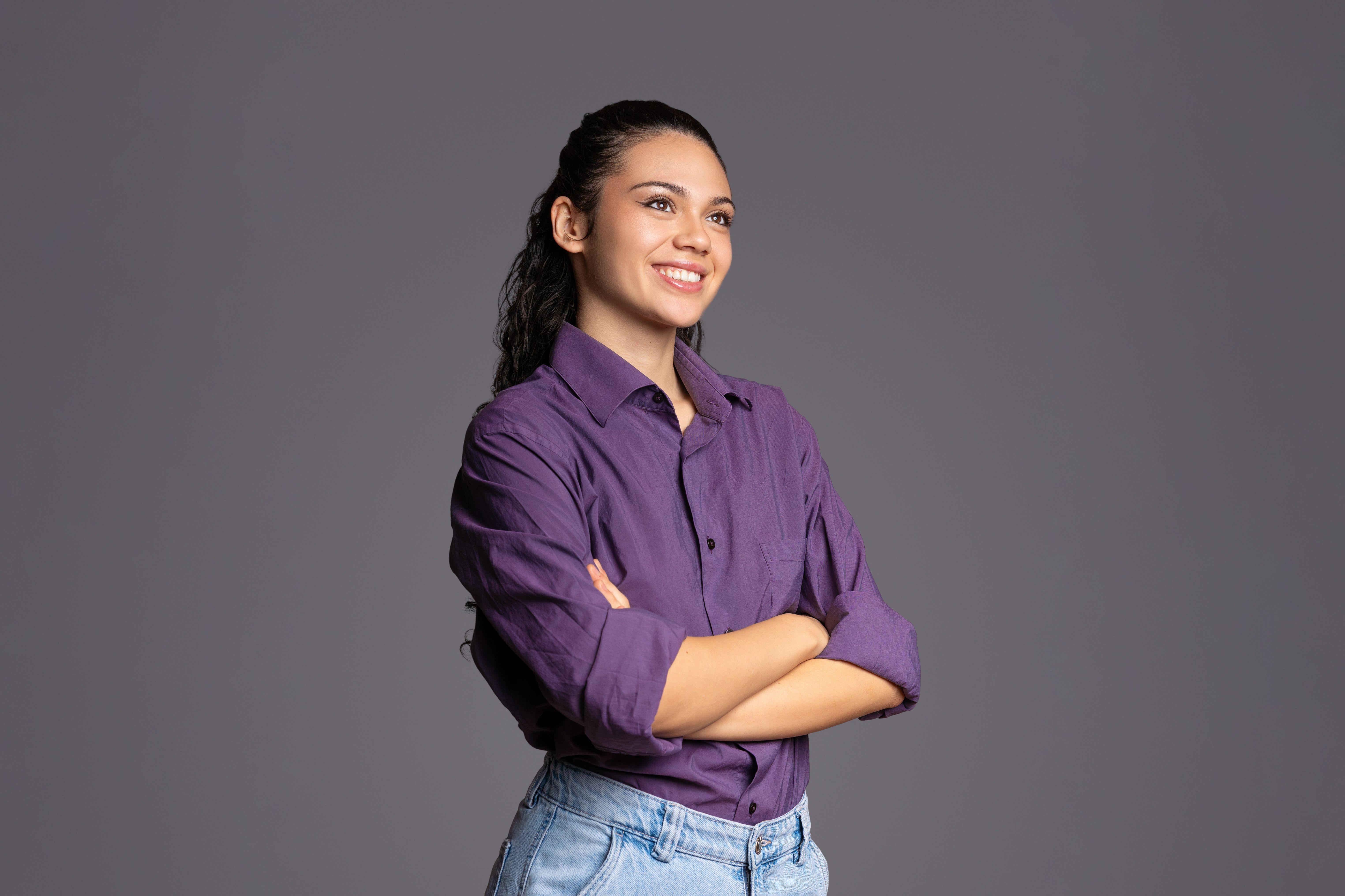 Confident latina professional crossing arms, turning head sideways with warm smile against neutral gray studio backdrop