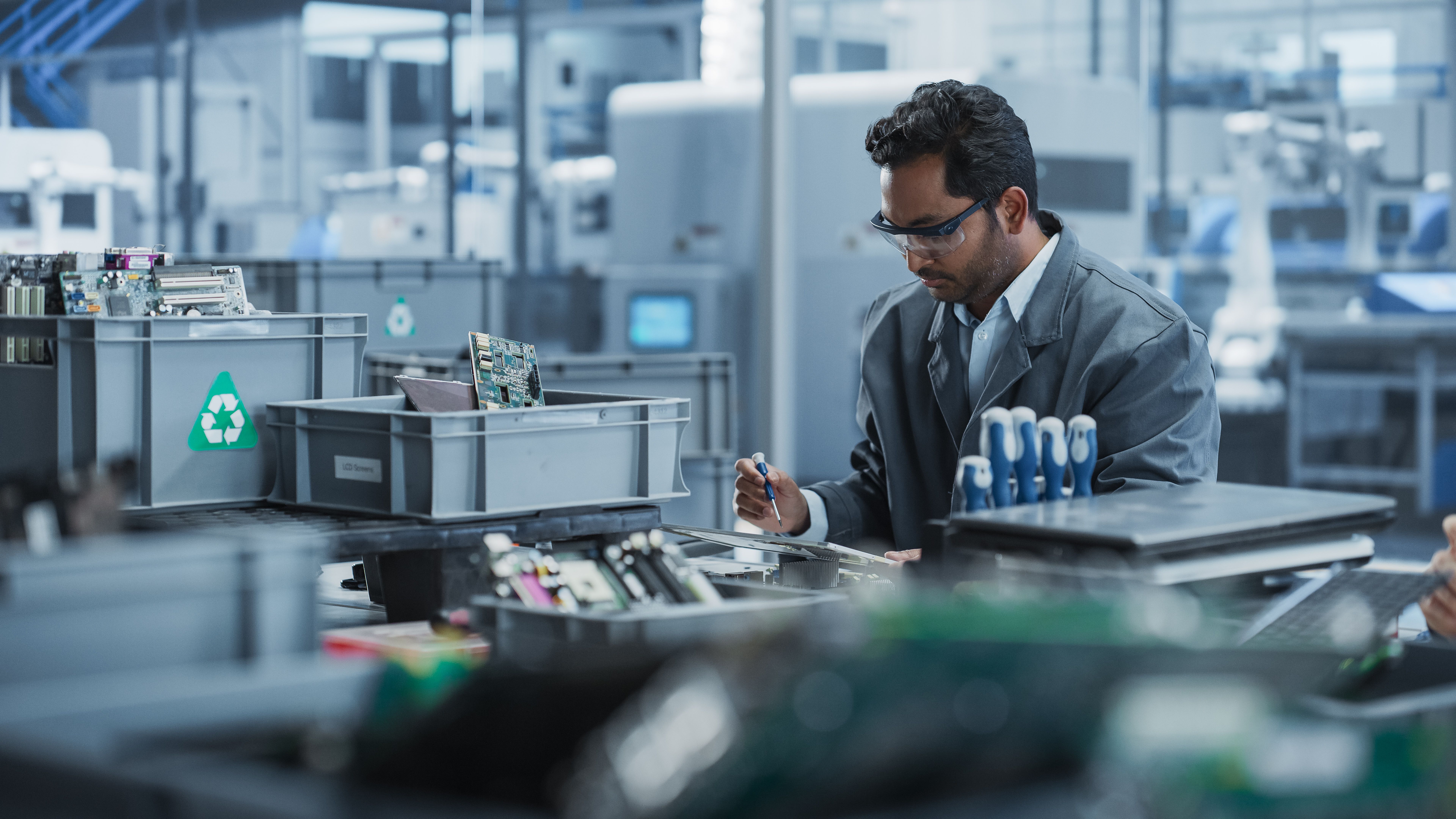 Indian Male Worker Disassembling Laptops To Recycle Electronic Components For Microchip Production At An Electronics Factory With Robotic Arms. Man Unscrewing Displays, Sorting Them Out