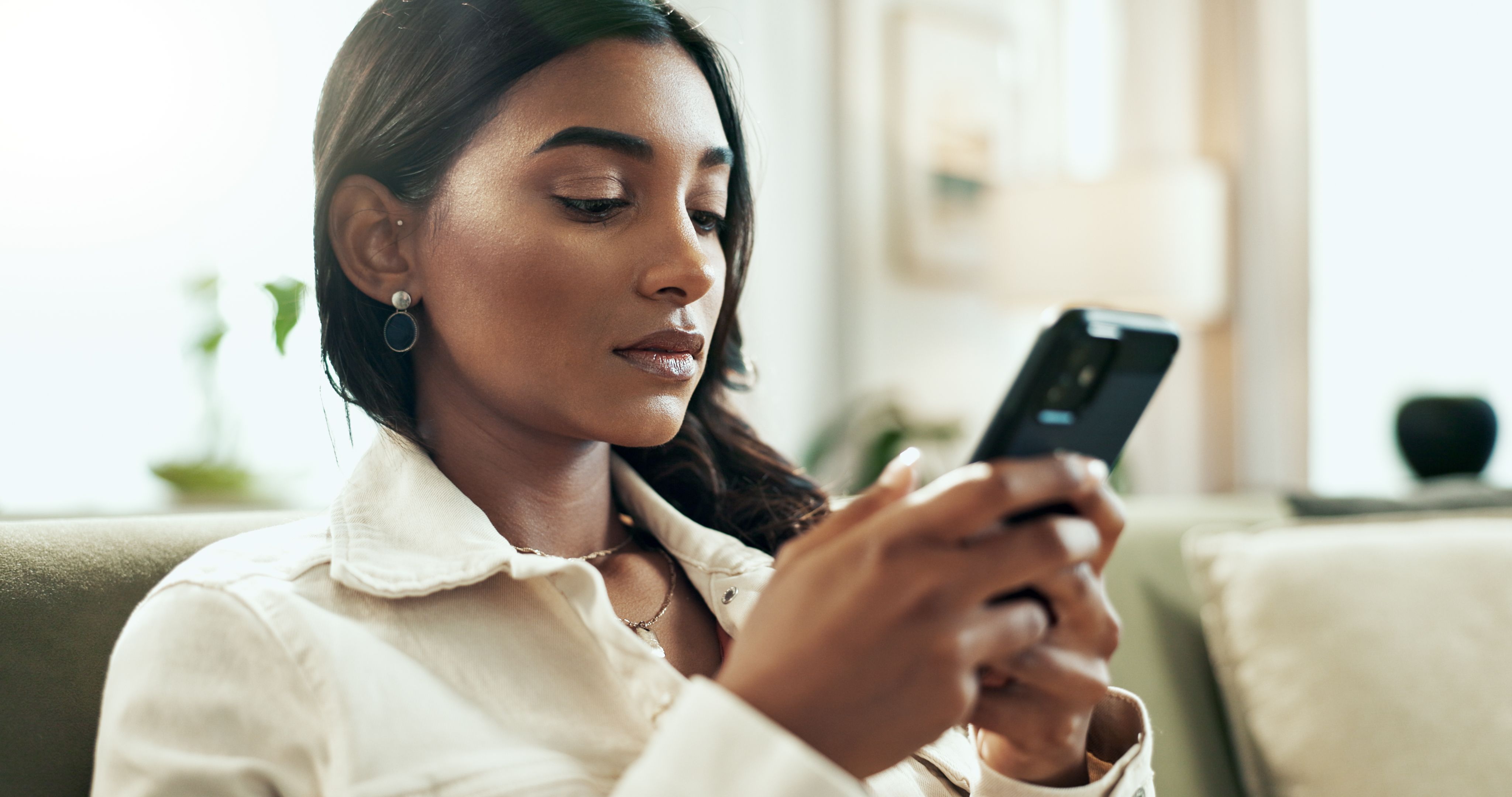 App, phone and serious Indian woman on sofa in living room of home for communication or research. Internet, social media and text message with person typing on mobile in apartment for networking