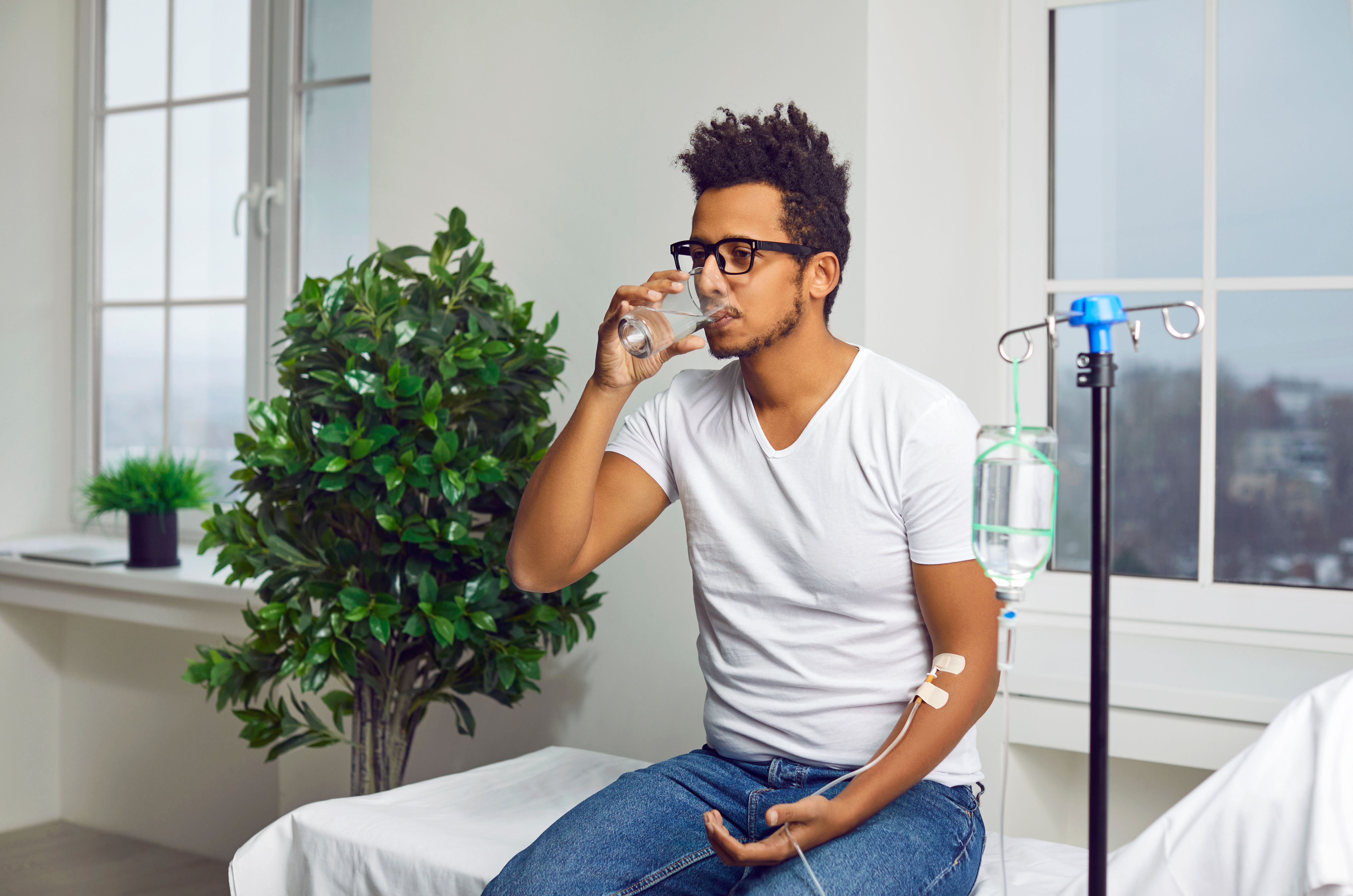 Young man drinks water while receiving vitamin therapy through IV drip line infusion