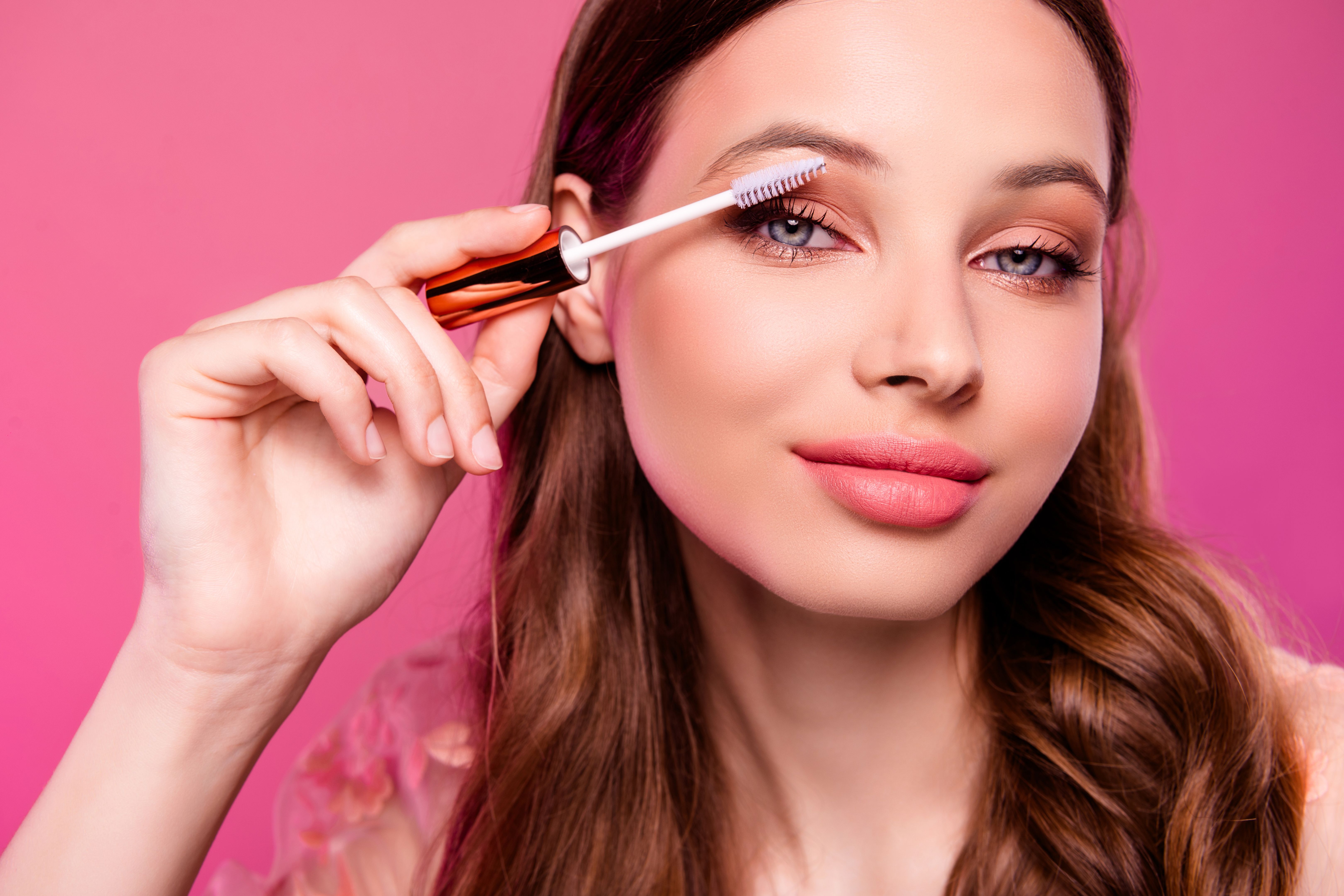 Close up photo of lovely stunning woman applying mascara for extra long lashes isolated on pink color background Close up photo of lovely stunning woman applying mascara for extra long lashes isolated on pink color background