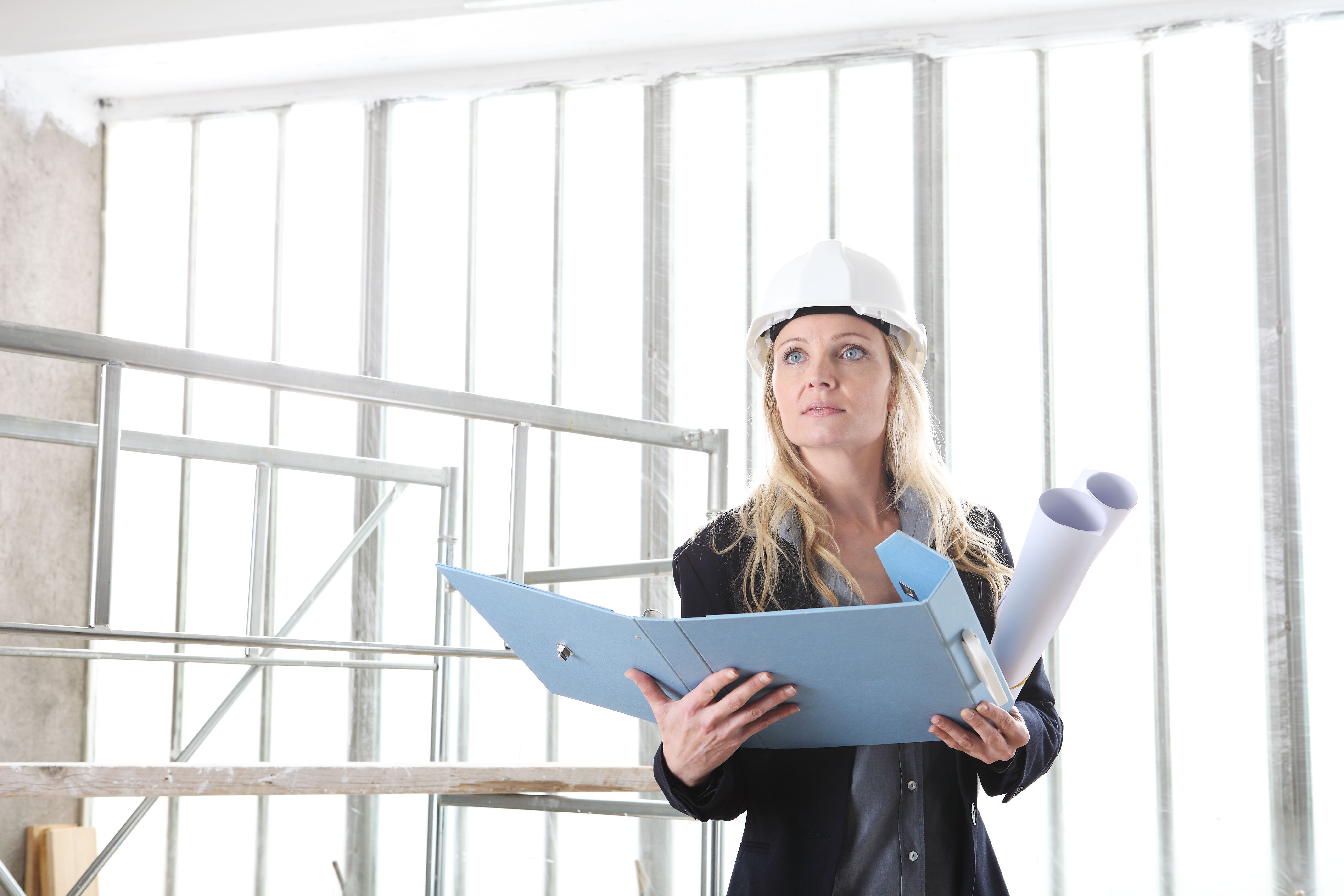 woman architect or construction engineer wear helmet and holds folder and blueprint inside a building site with windows and scaffolding in the background with copy space woman architect or construction engineer wear helmet and holds folder and blueprint inside a building site with windows and scaffolding in the background with copy space