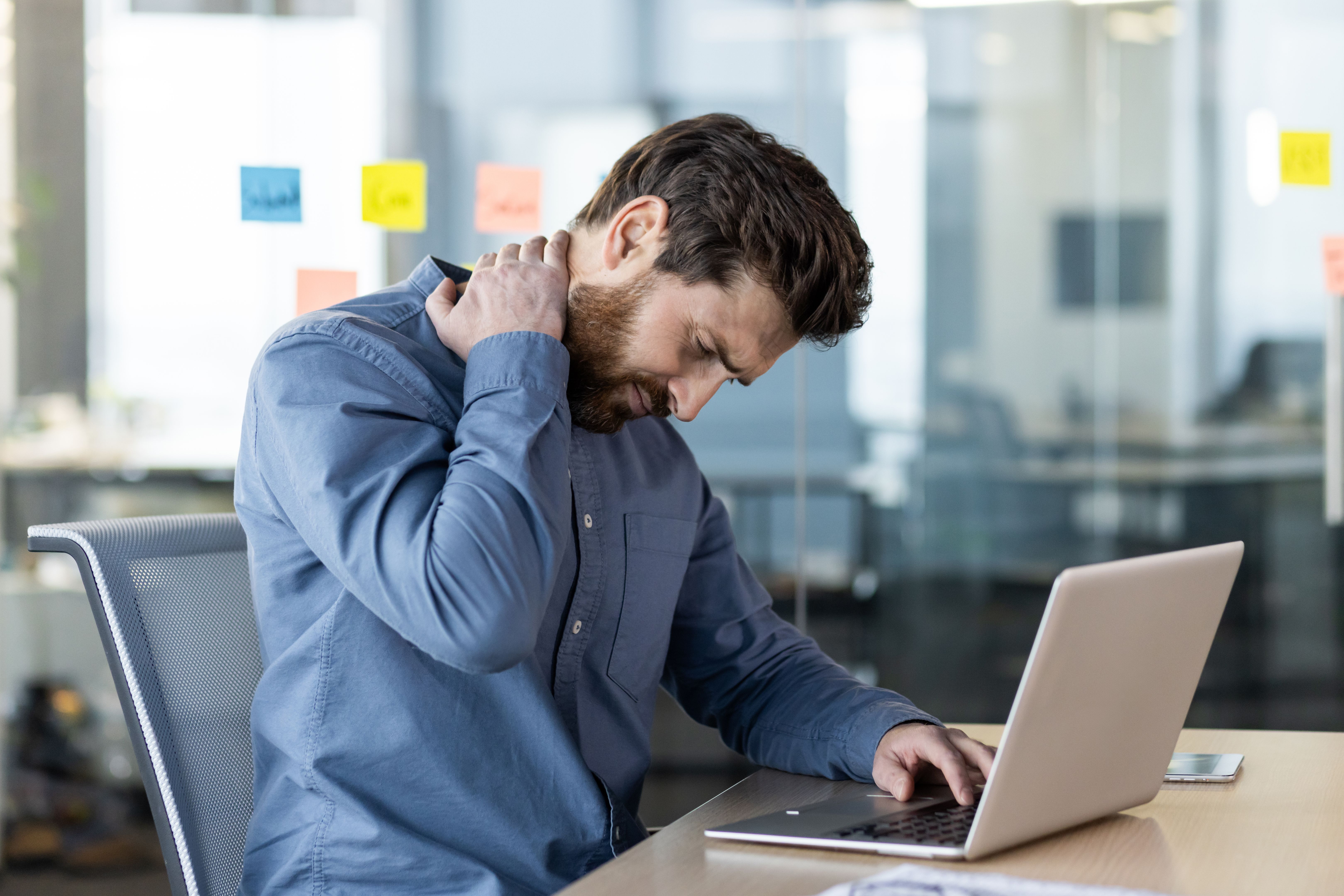 A tired young man in a blue shirt sits in the office at a desk, works on a laptop and massages his neck with his hand