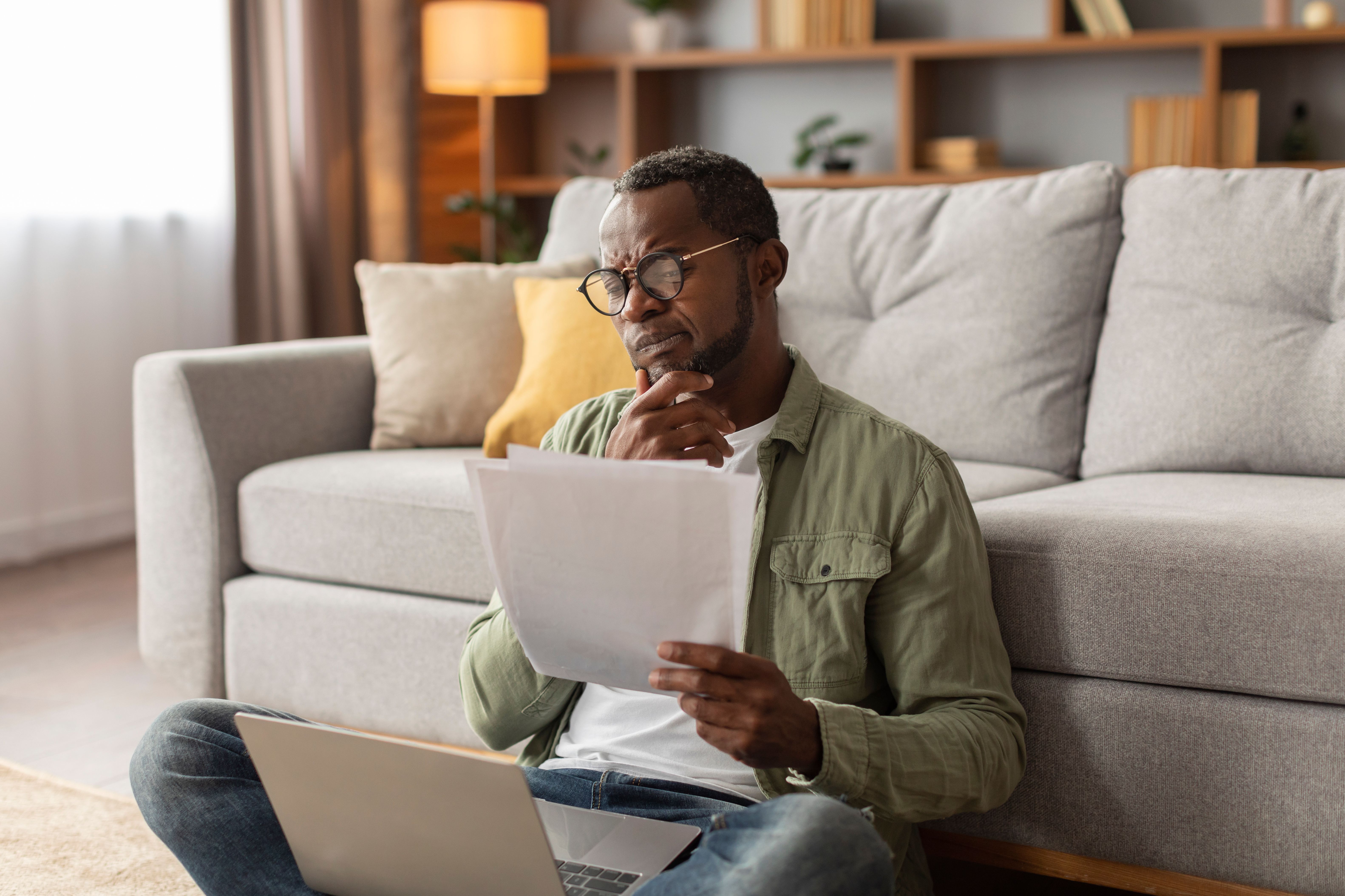 Pensive serious adult black man in glasses uses computer, works with documents, sits on floor in living room Pensive serious adult black man in glasses uses computer, works with documents, sits on floor in living room