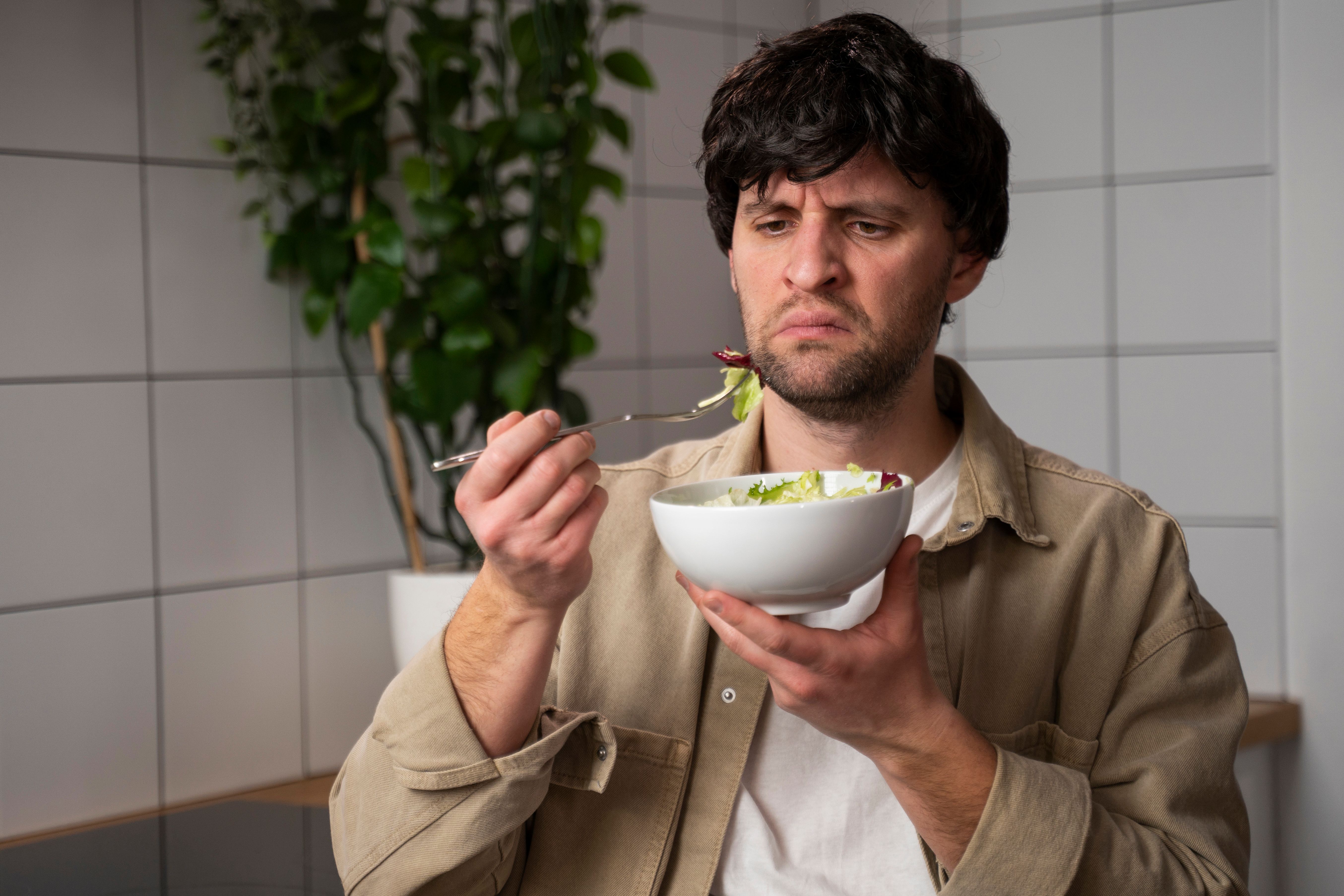 A man in a beige shirt eats a tasteless salad in the kitchen