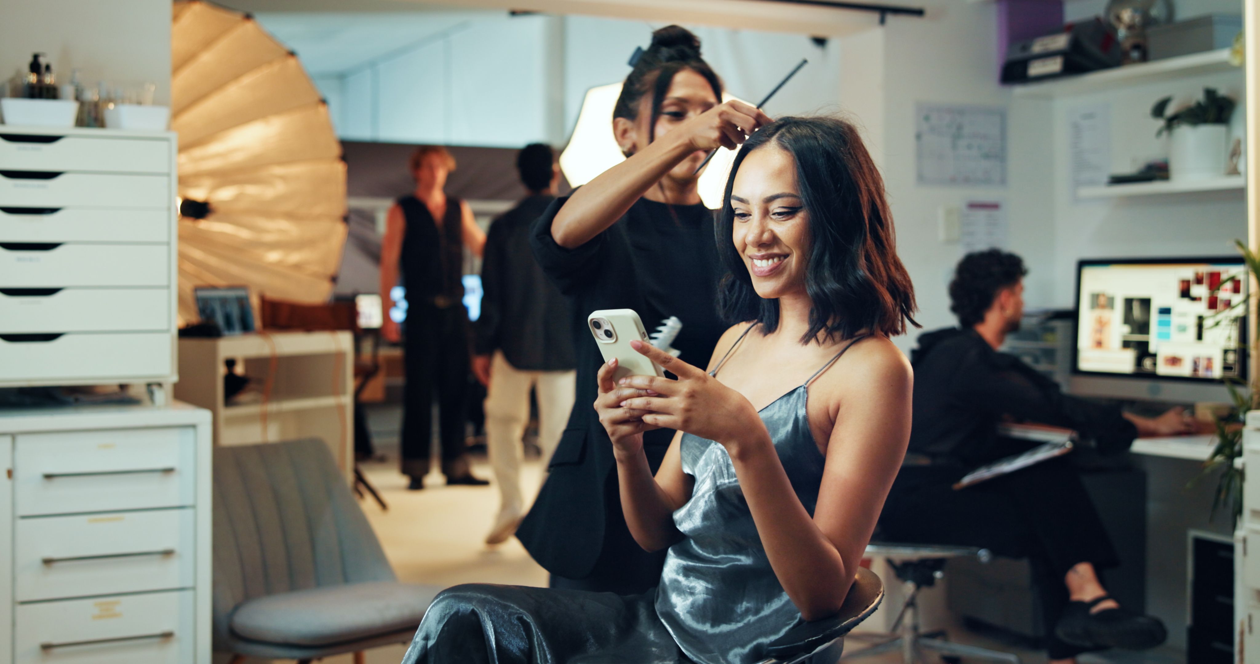 Makeup, phone or photography with hair stylist and model in studio for behind the scenes preparation. App, getting ready and social media with woman on set for catalog or magazine photoshoot