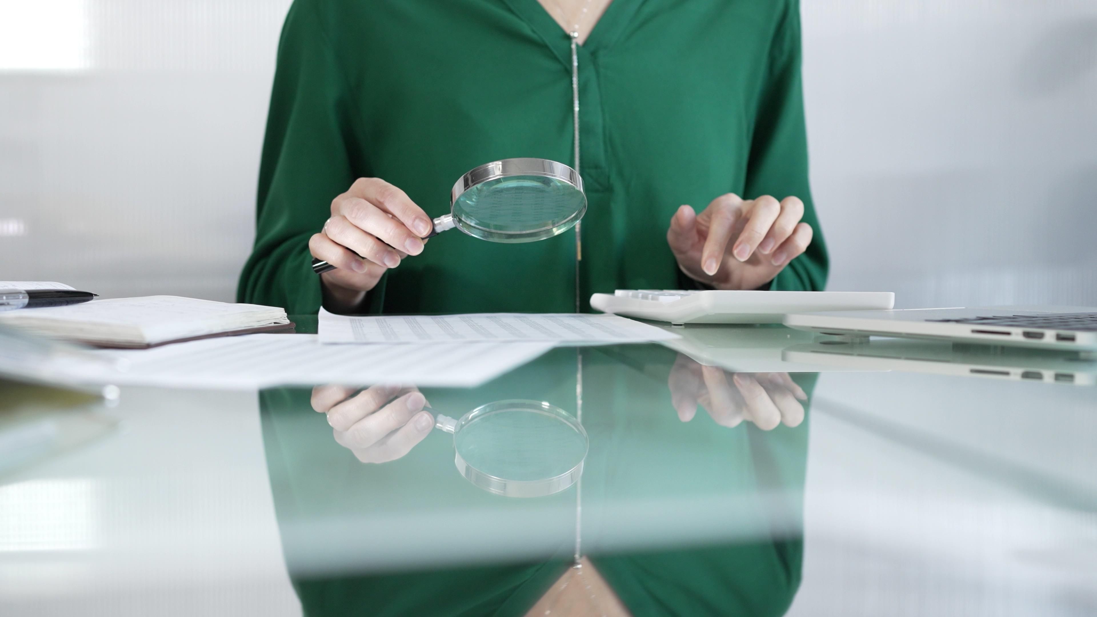 Businesswoman in green blouse carefully reviewing financial reports with magnifying glass and calculator, conducting audit and analyzing data in office. Taxes, audit in business