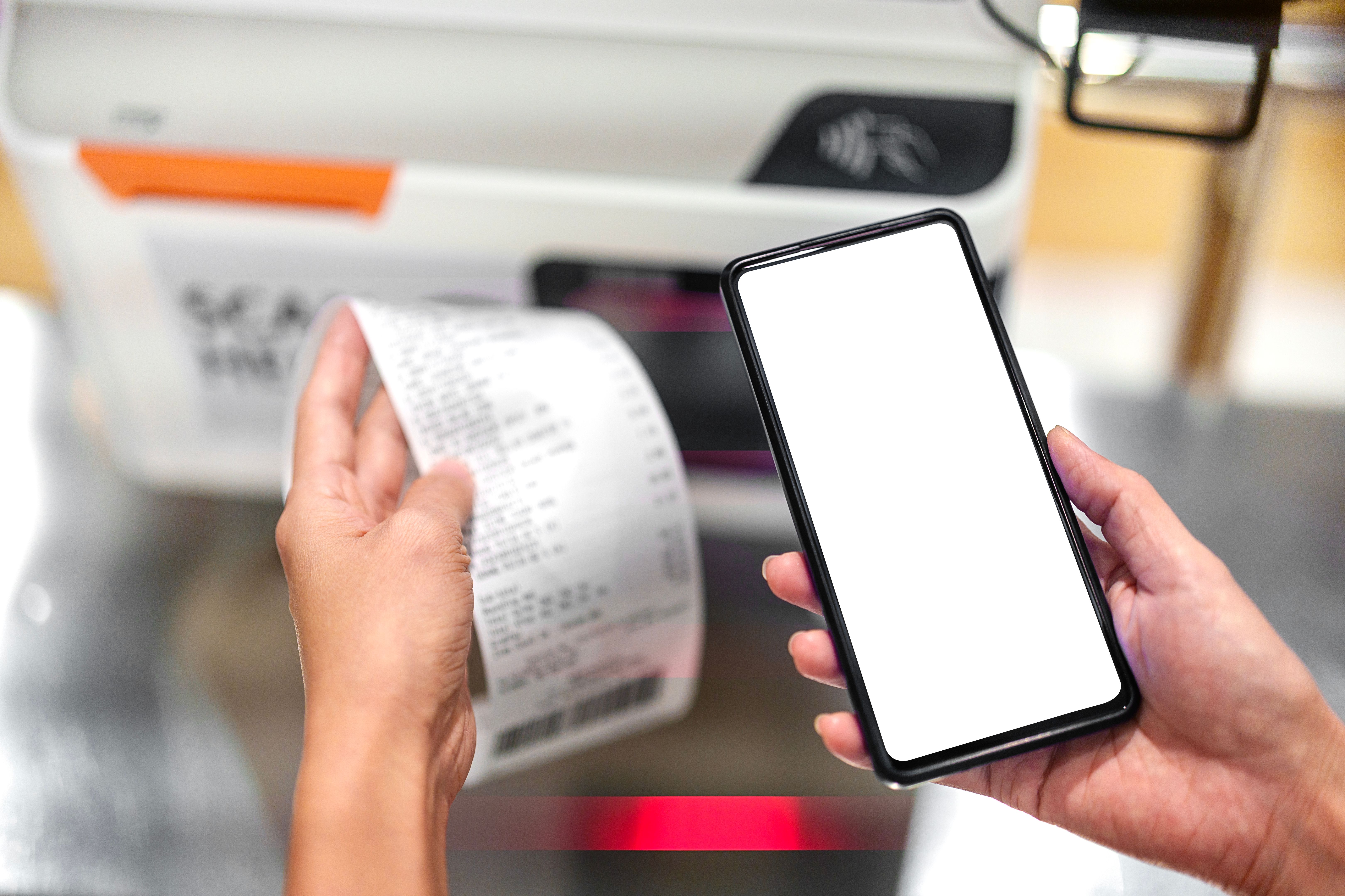 Close up image of an Asian woman is seen checking her grocery receipt while holding a smartphone with a blank screen at the grocery store's self-checkout service. Close up image of an Asian woman is seen checking her grocery receipt while holding a smartphone with a blank screen at the grocery store's self-checkout service.