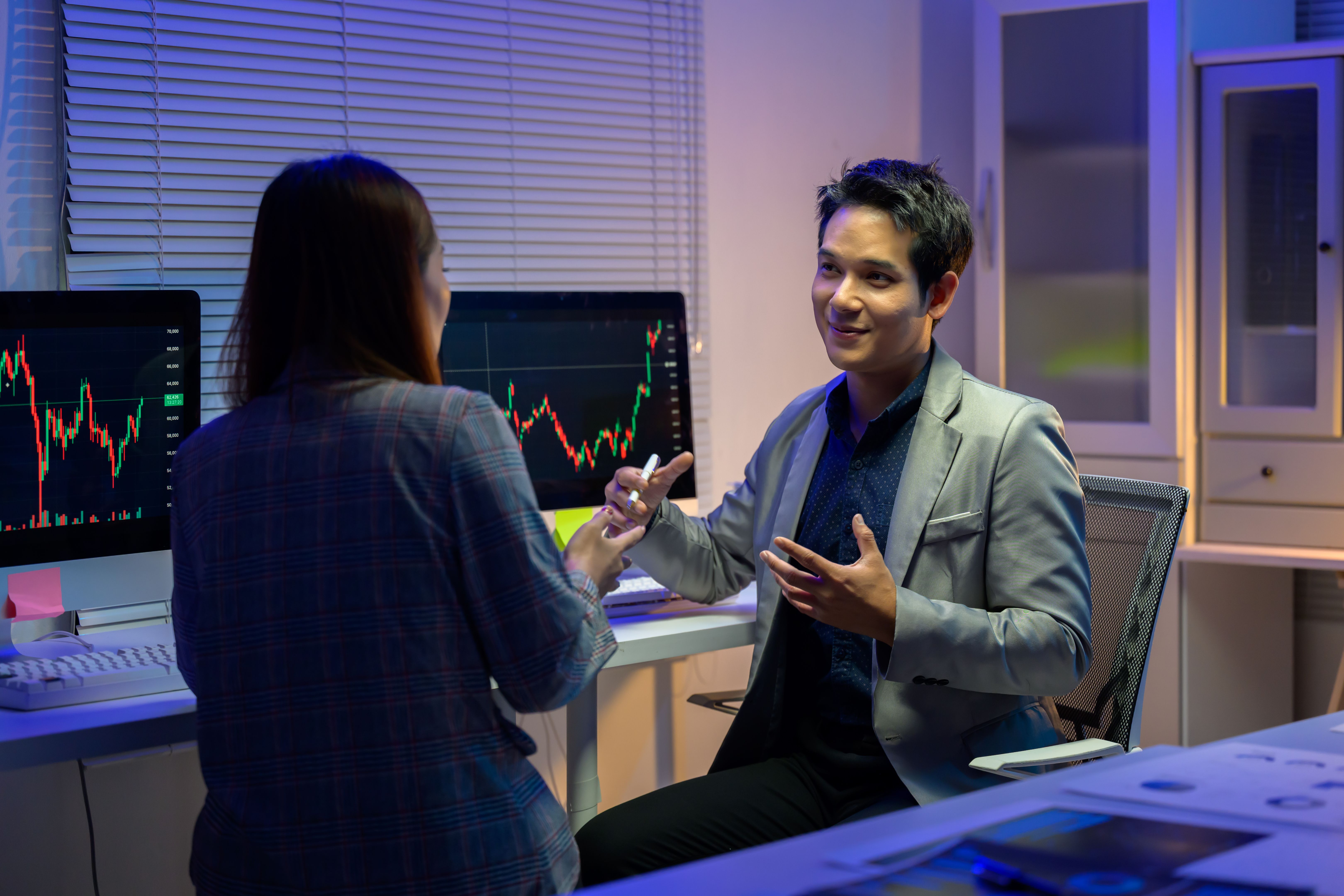 Two stockbrokers are discussing candlestick charts displayed on computer screens, analyzing market trends and investment strategies in their office at night