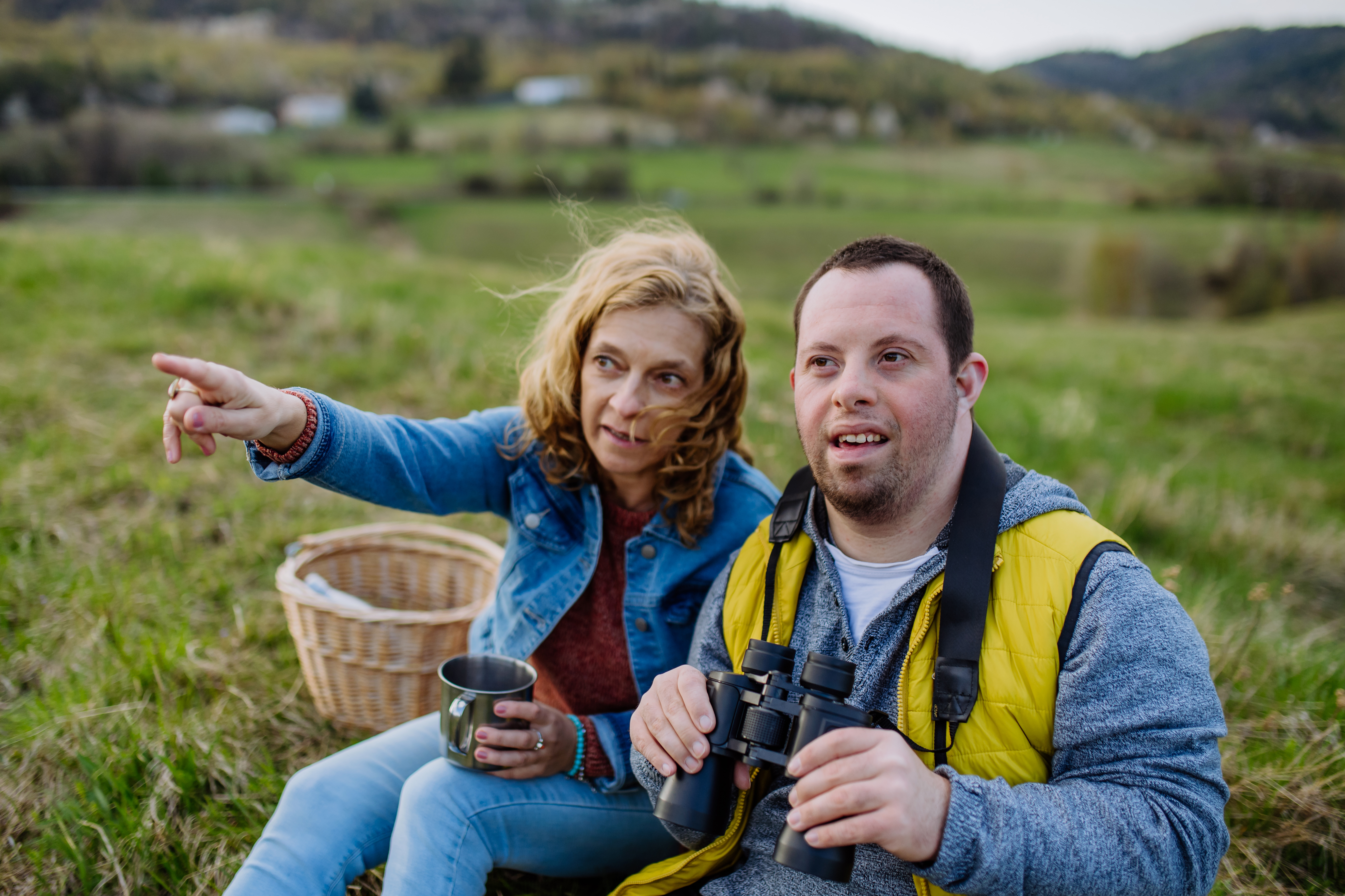 Portrait of happy young man with Down syndrome with his mother resting in nature, sitting and looking at view with binoculars.