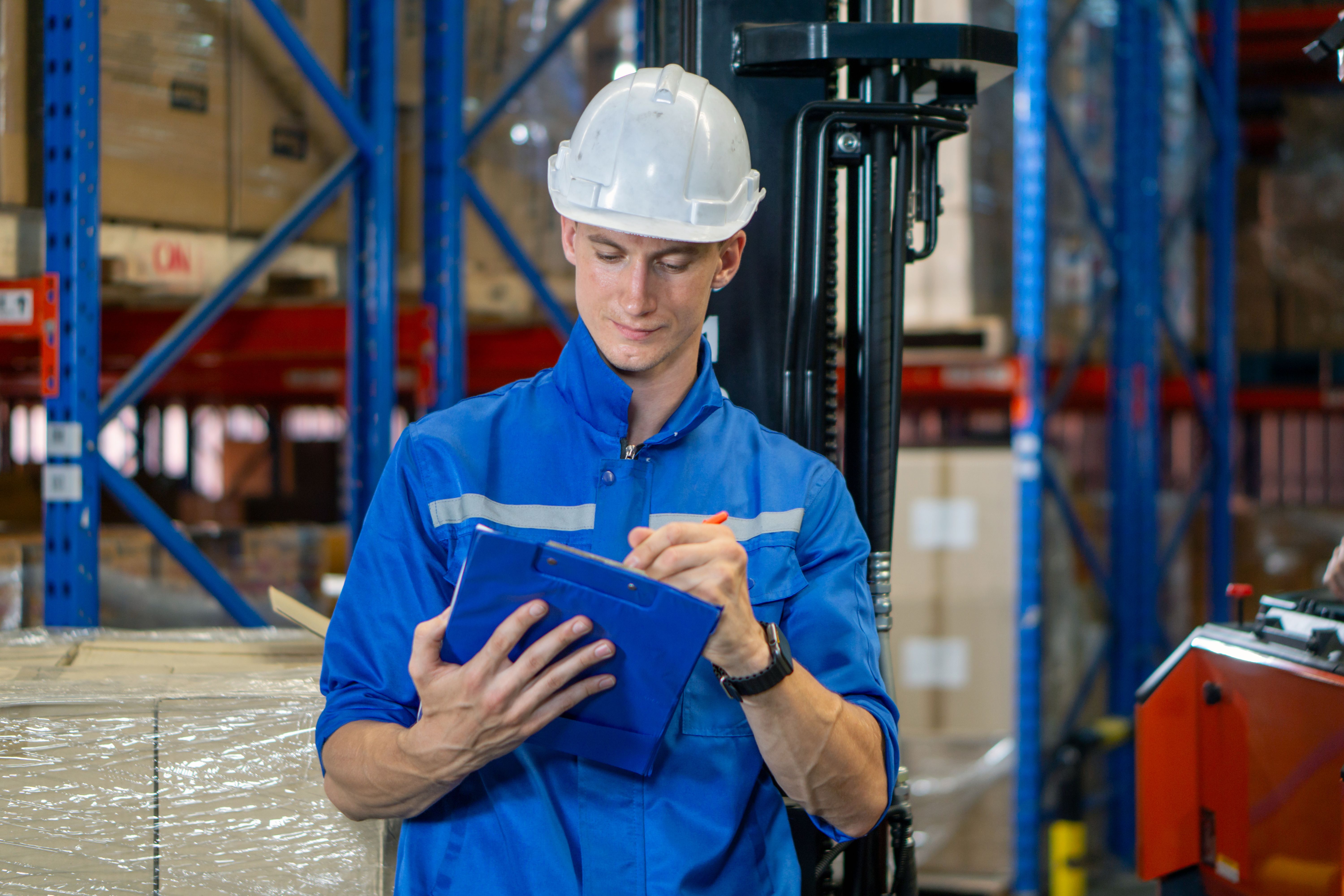 Giovane lavoratore in uniforme blu e casco di sicurezza esamina il record dell'inventario sugli appunti. Mettiti tra le scatole impilate in un'area di stoccaggio affollata. Atmosfera di lavoro nel magazzino generale con sistema di scaffalature