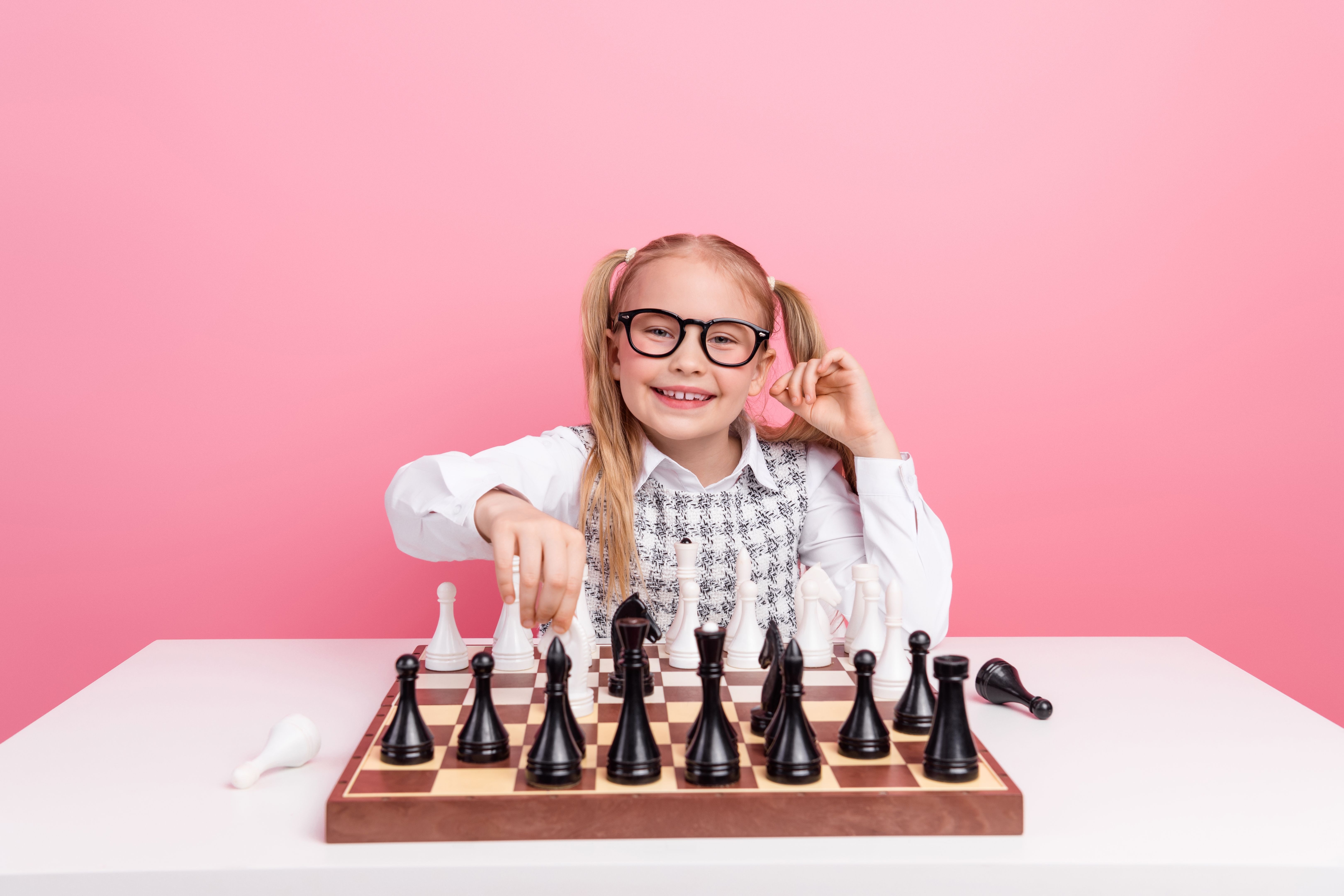 Cheerful schoolgirl learning chess strategies on a pink background, emphasizing intelligence, learning, and positive energy