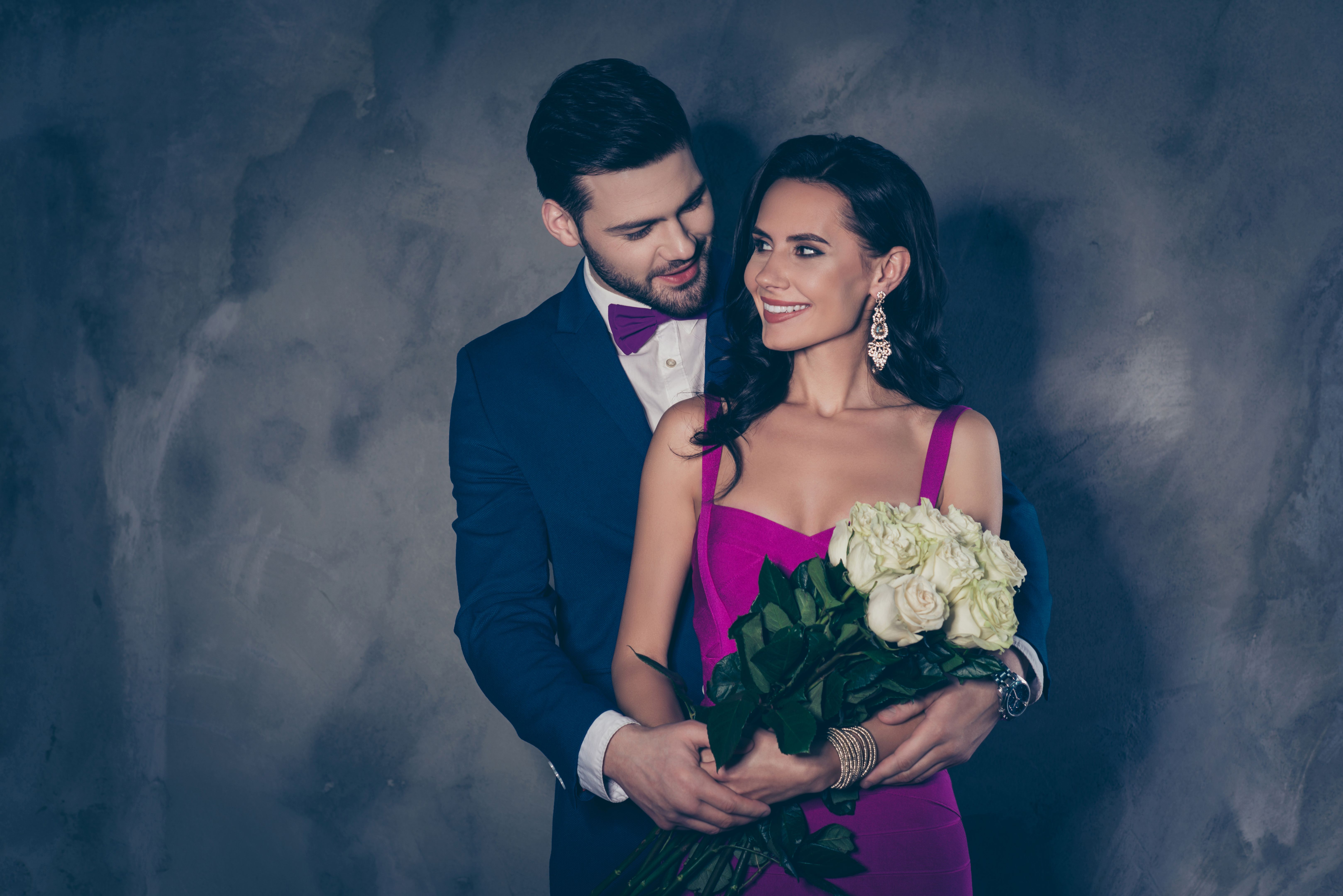 Portrait of lady in purple dress with jewelry gentleman in tuxedo with bowtie holding white roses in hands, lovely romantic spouses isolated on grey background looking at each other