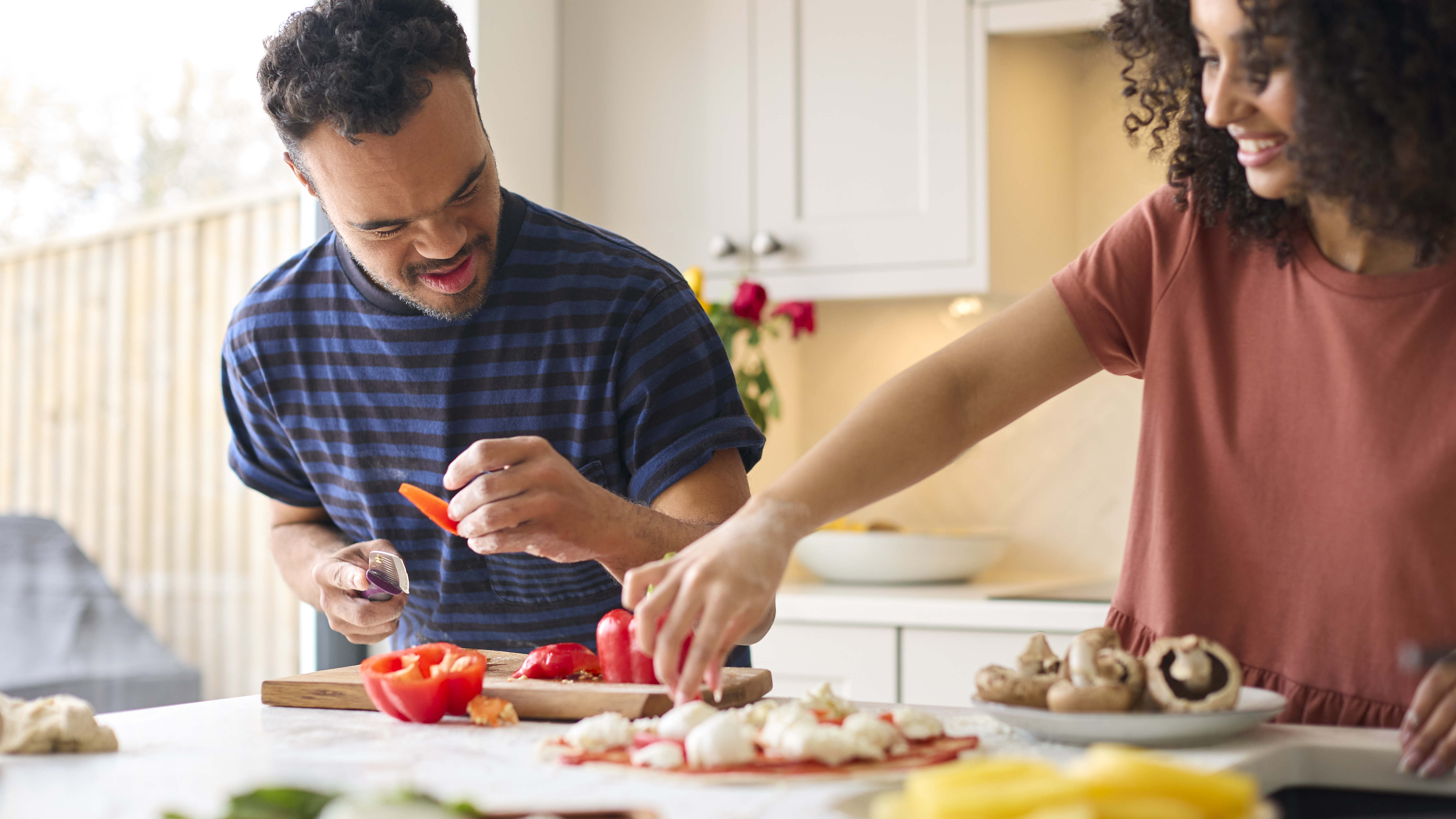 Couple At Home With Man With Down Syndrome And Woman Preparing Topping For Pizza In Kitchen Together