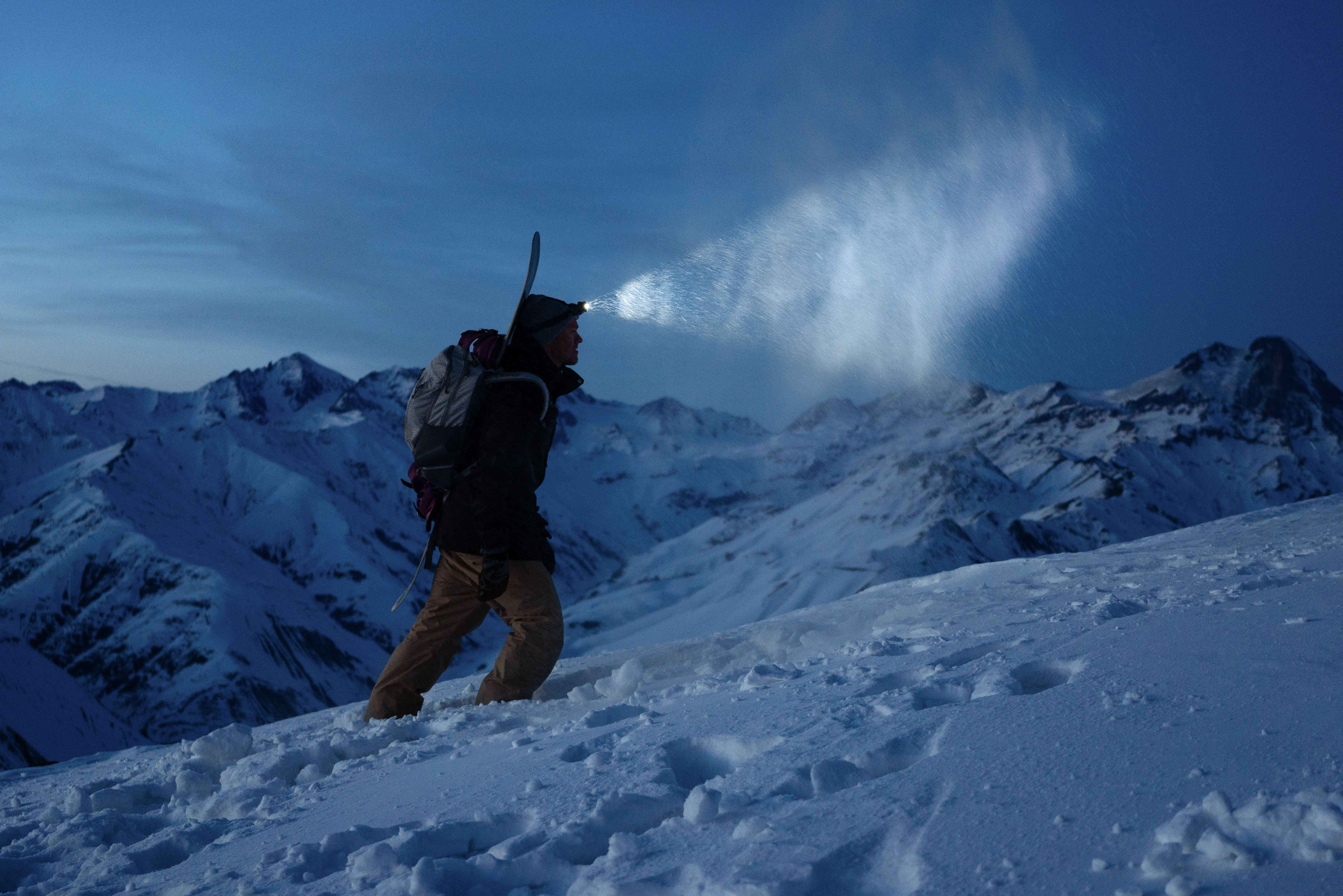 Ski touring man commit climb on night winter mountain. Tourist with headlamp, backpack and a snowboard behind his back walking on snowy slope. Backcountry