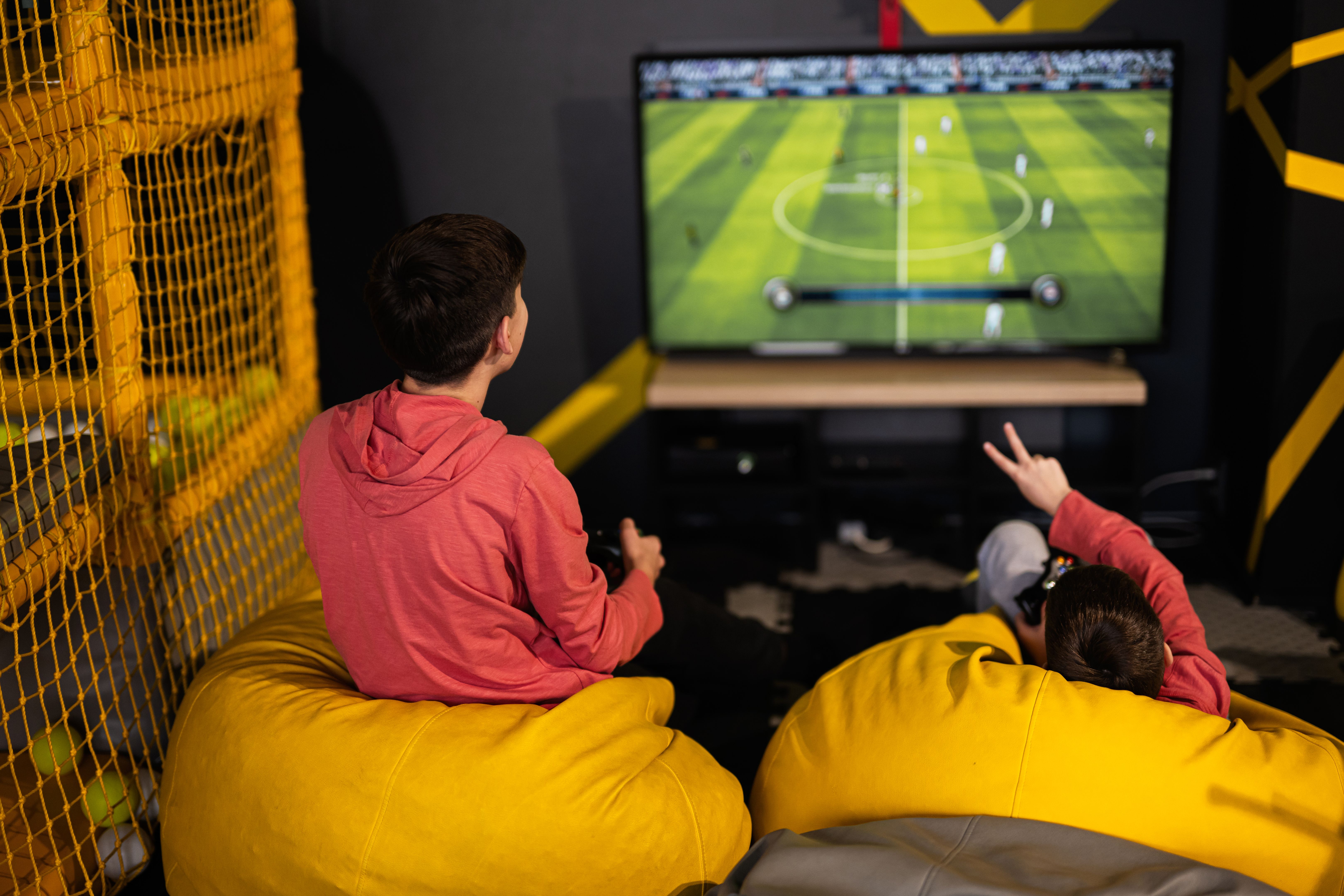 Two brothers playing football video game console, sitting on yellow pouf in kids play center. Two brothers playing football video game console, sitting on yellow pouf in kids play center.