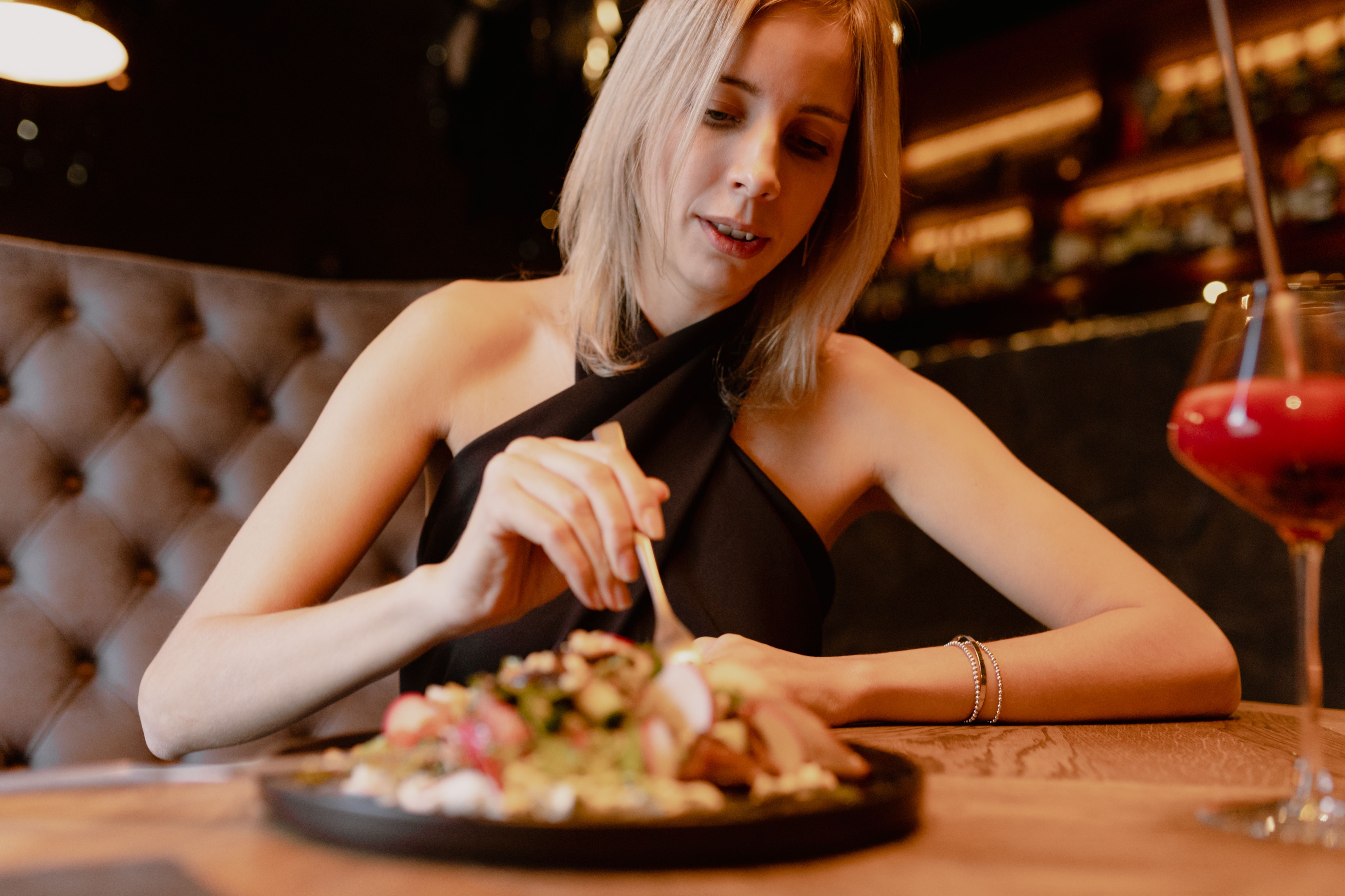 Portrait of young focused woman sitting on brown sofa near black plate with vegetable salad and glass with red cocktail. Portrait of young focused woman sitting on brown sofa near black plate with vegetable salad and glass with red cocktail.