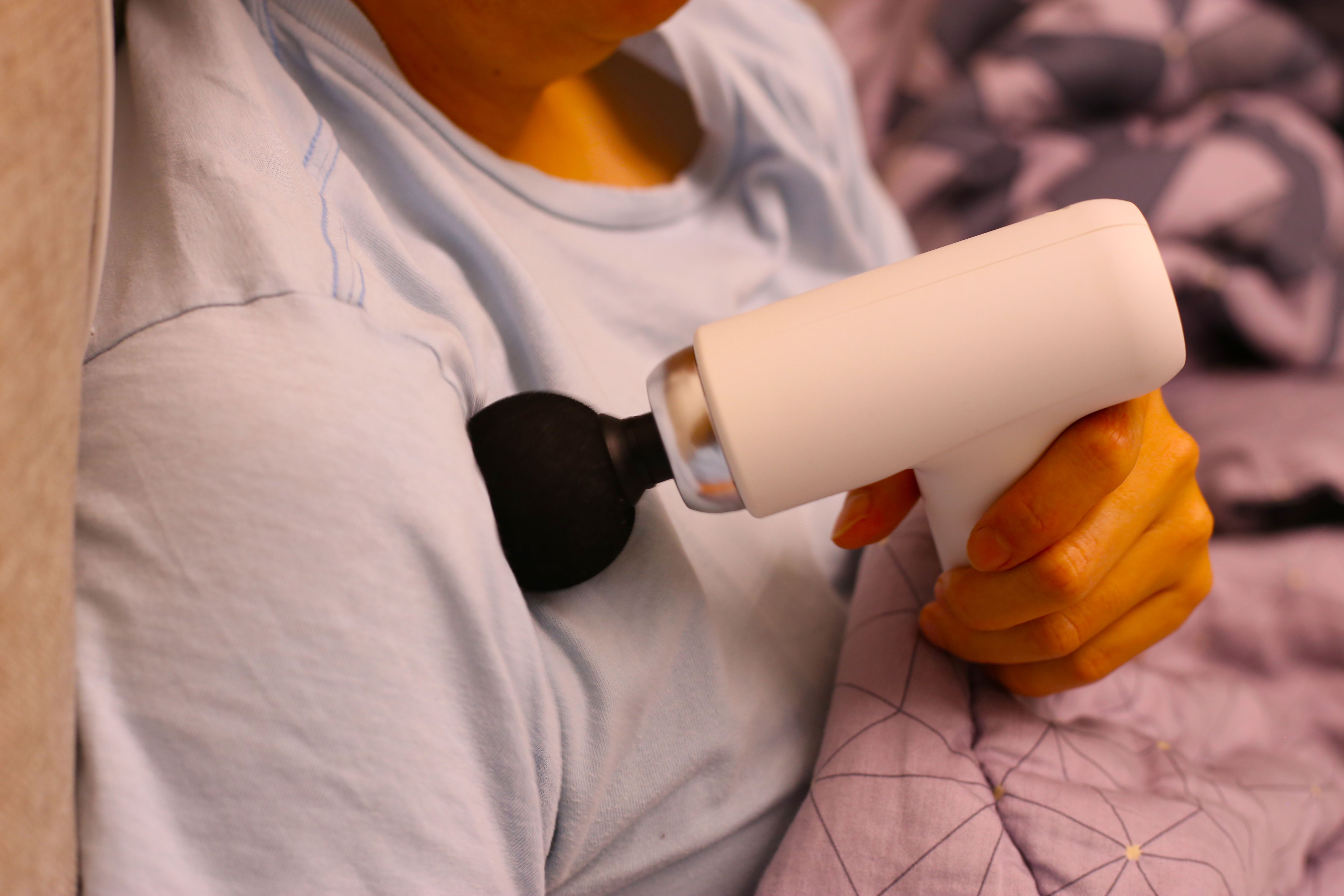 Person using a handheld massage gun on their shoulder while sitting on a couch. Person using a handheld massage gun on their shoulder while sitting on a couch.