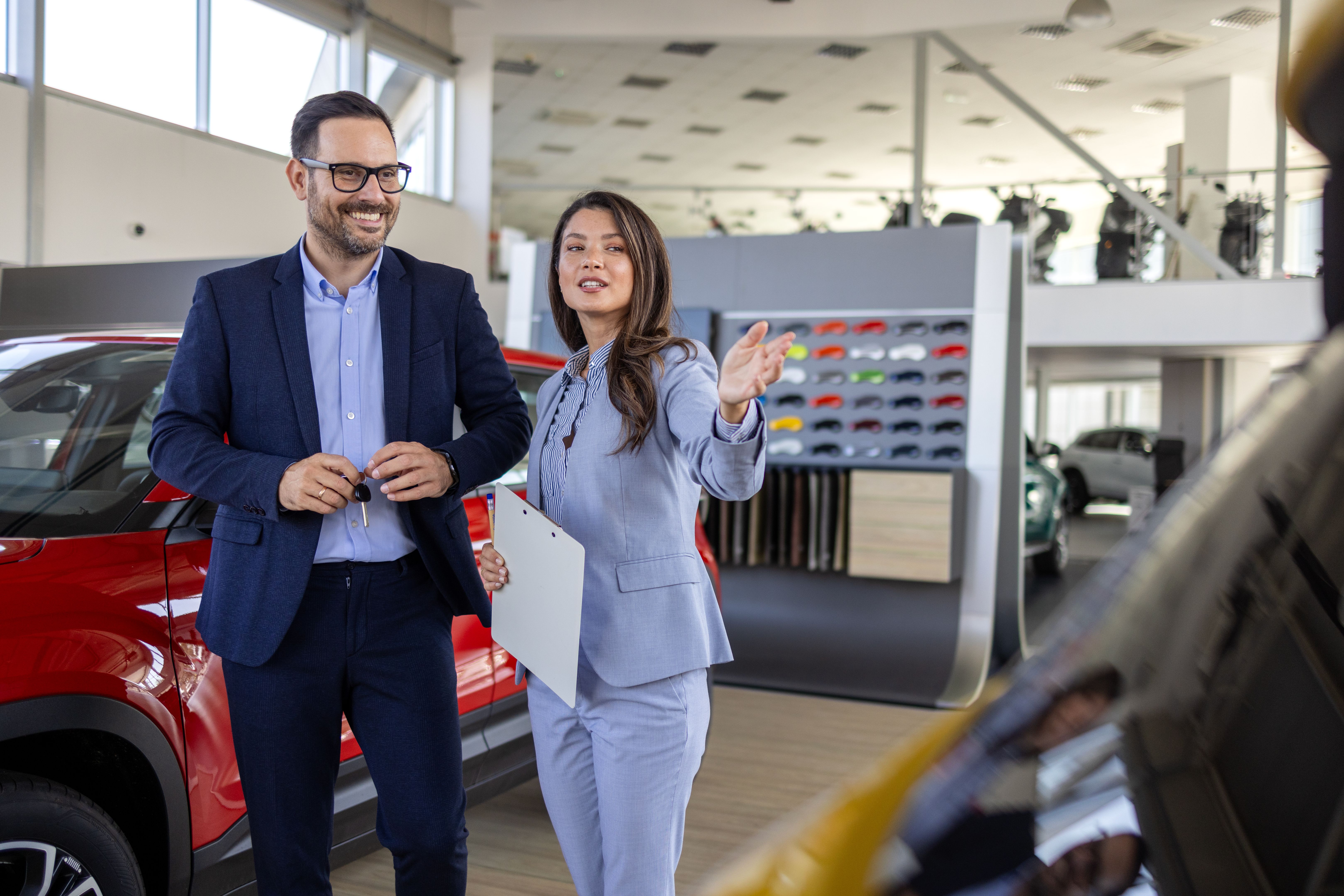 Super sales team in dealership, two consultants or managers in elegant suits with laptop and tablet in arms in car dealership