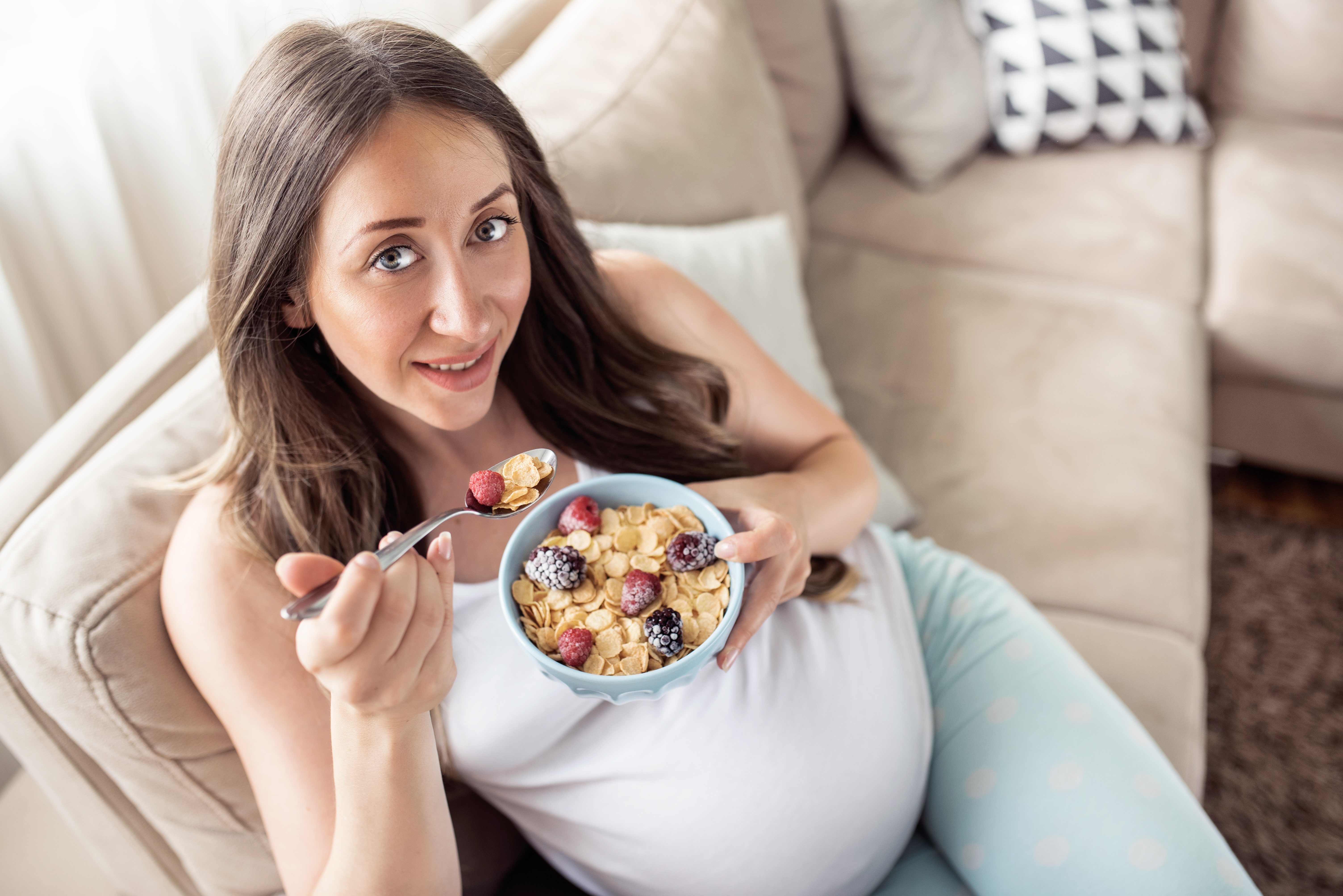 Top view of happy pregnant woman eating cereals with fruits for breakfast in bed at home. Looking at camera.