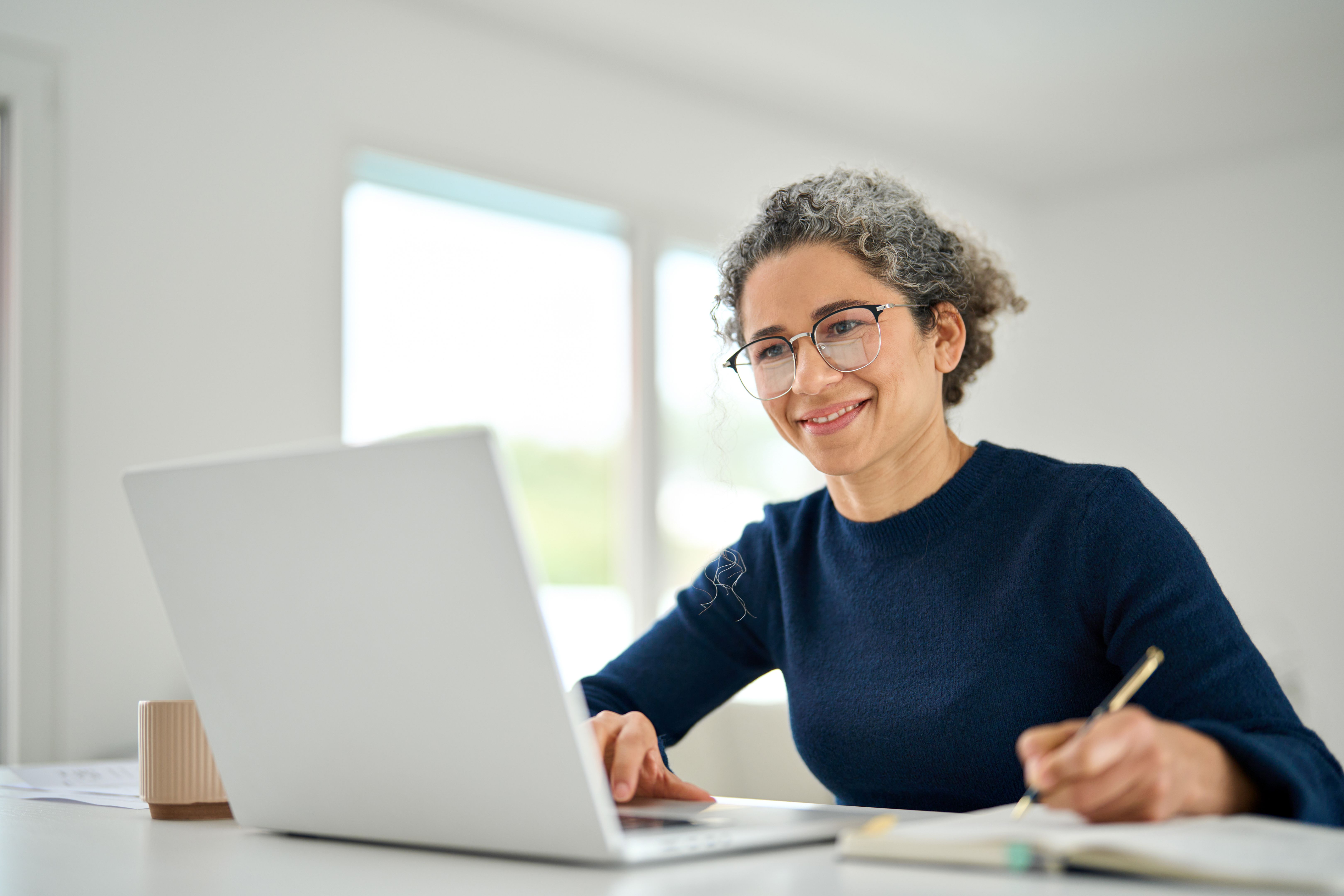 Happy middle aged woman sitting at table and using laptop writing notes.