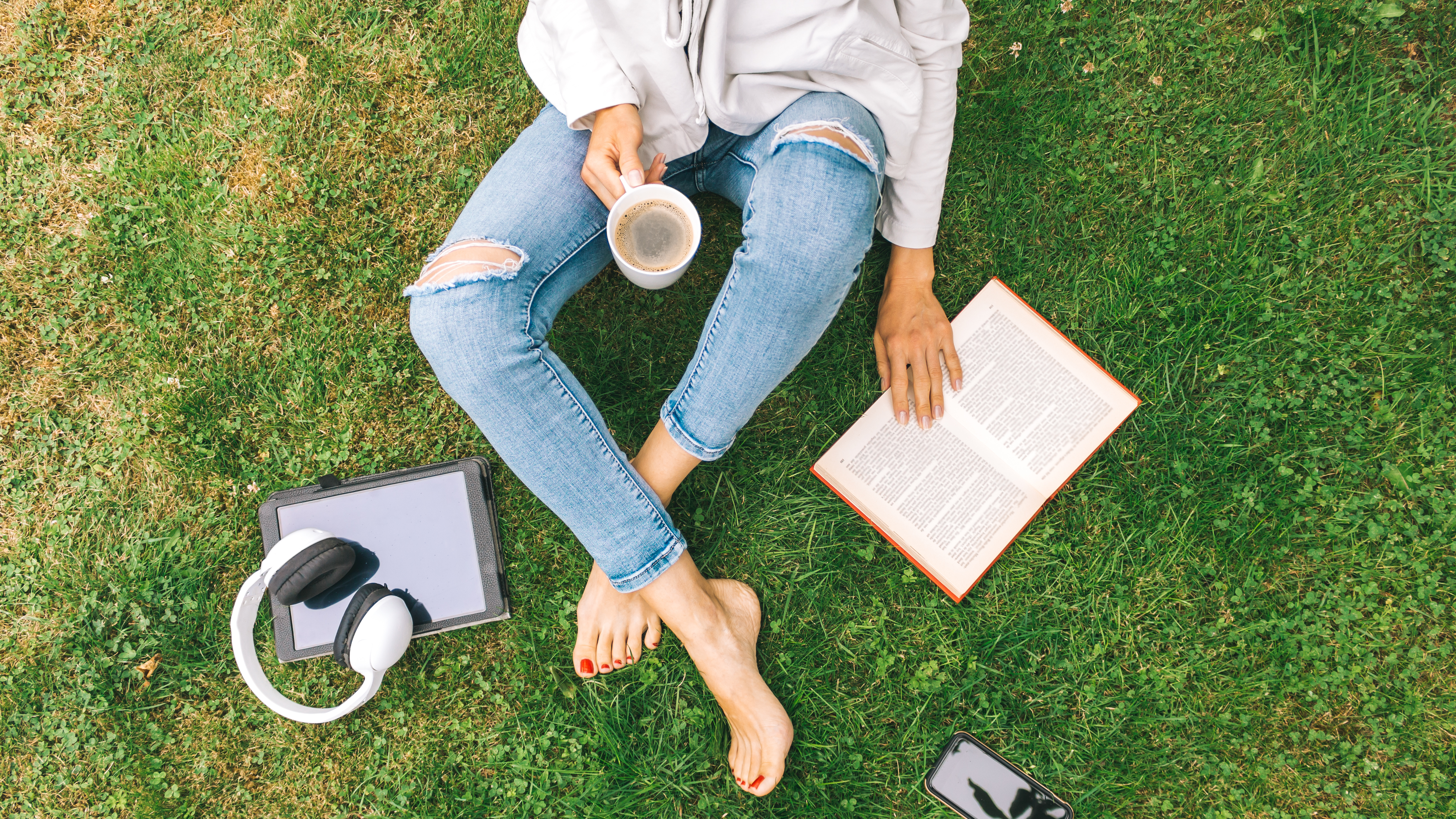 Young woman sitting on the grass drinking coffee and reading a book enjoys outdoor recreation. Young woman sitting on the grass drinking coffee and reading a book enjoys outdoor recreation.