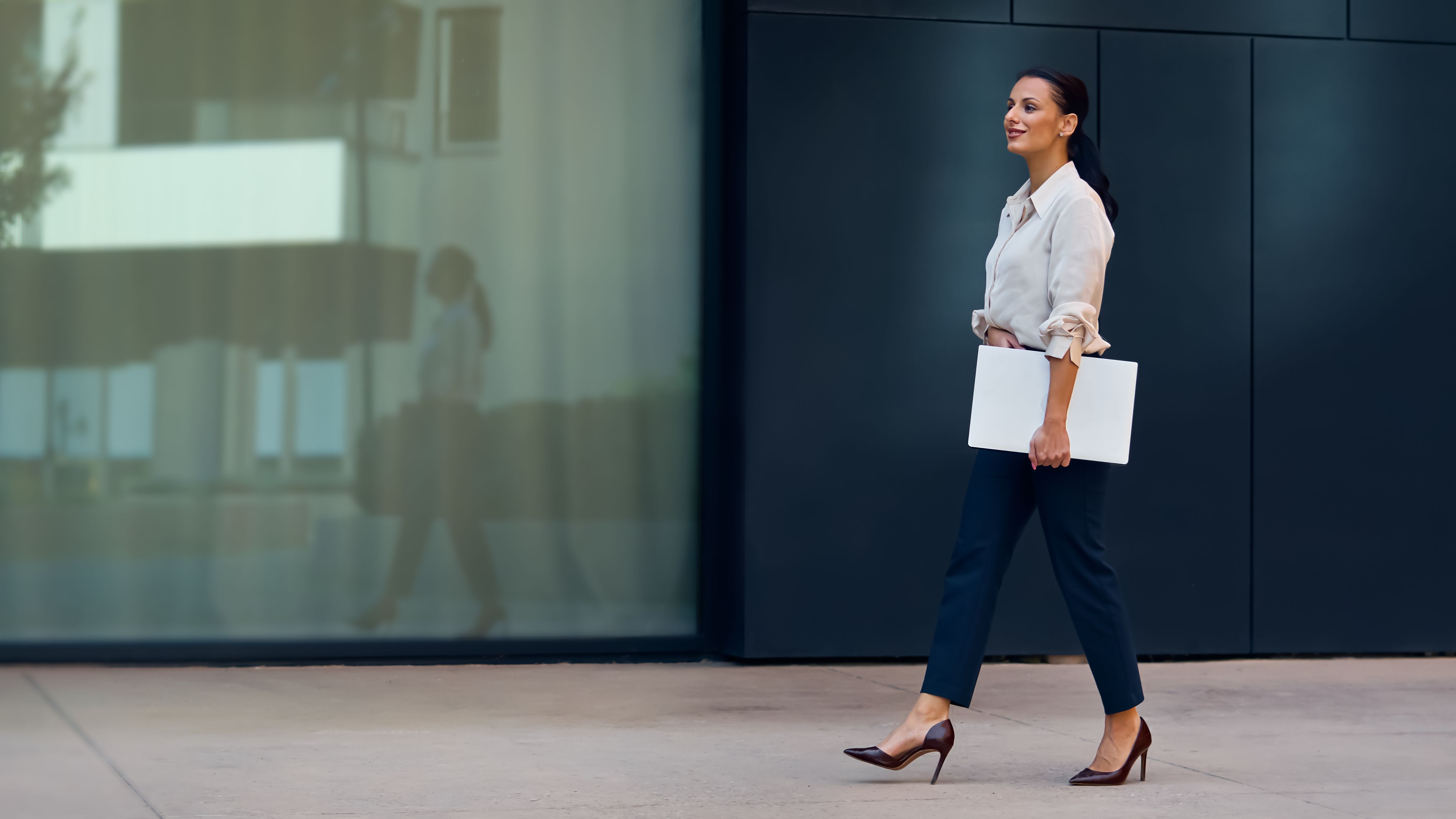 Professional Woman Walking Outdoors With Laptop Near Modern Office Building, Confident and Focused