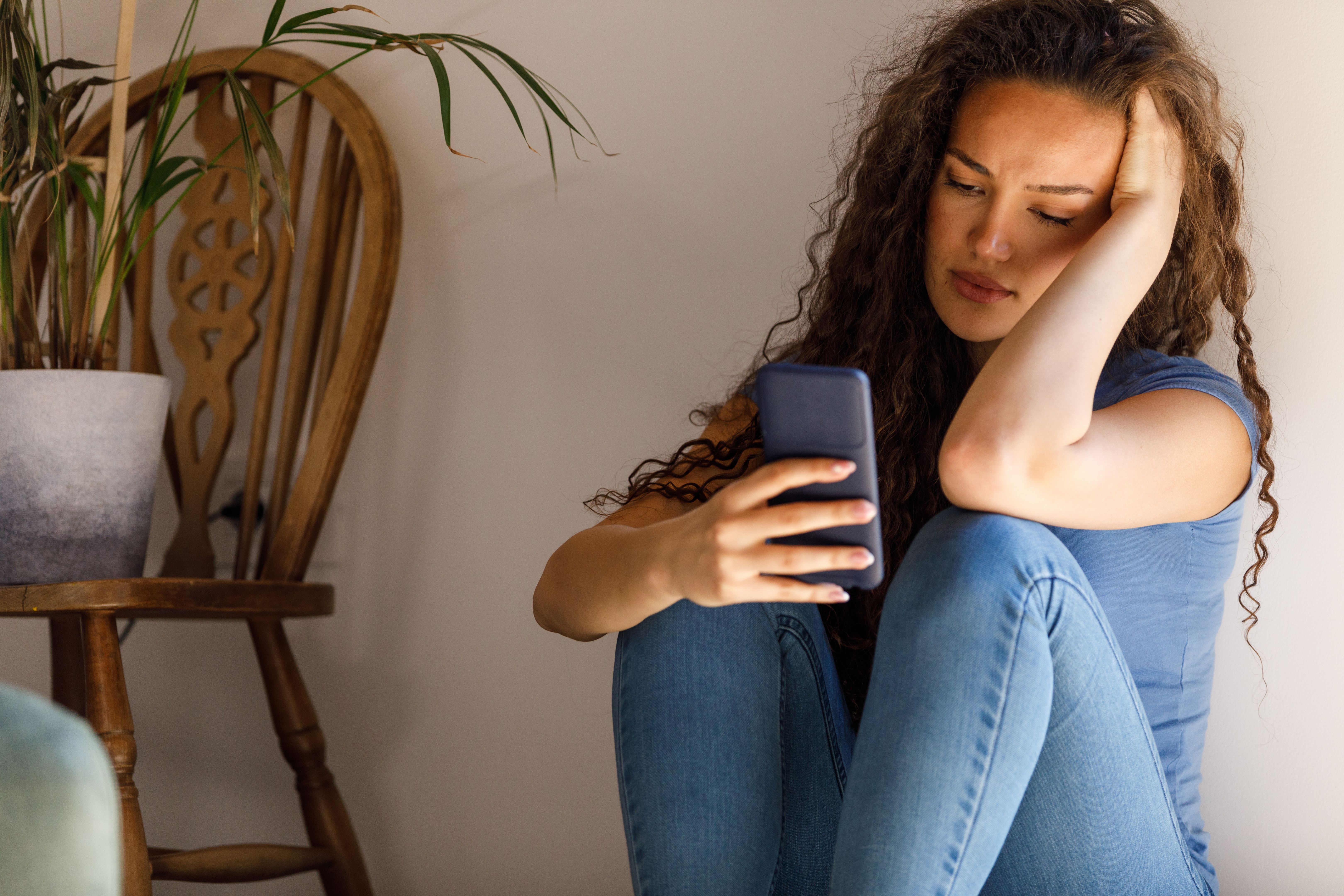 Young woman sitting on the floor, against the wall, responding to a text message that is causing her anxiety