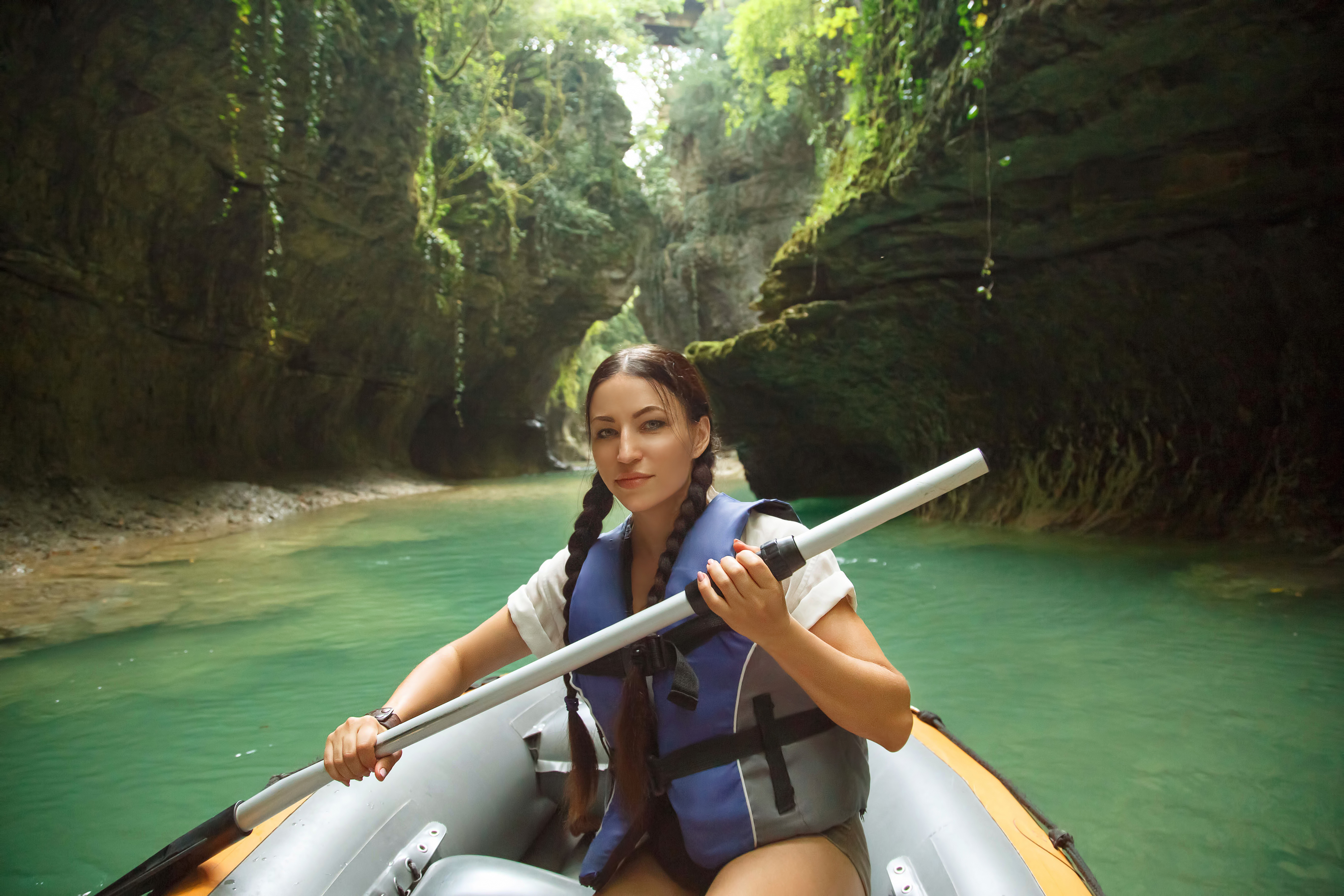 pretty young woman with dark hair gathered in two black braids, lady looks and poses for camera in life jacket and paddle in hands, tourist in inflatable boat sails through Martvili Kanyon Georgia pretty young woman with dark hair gathered in two black braids, lady looks and poses for camera in life jacket and paddle in hands, tourist in inflatable boat sails through Martvili Kanyon Georgia