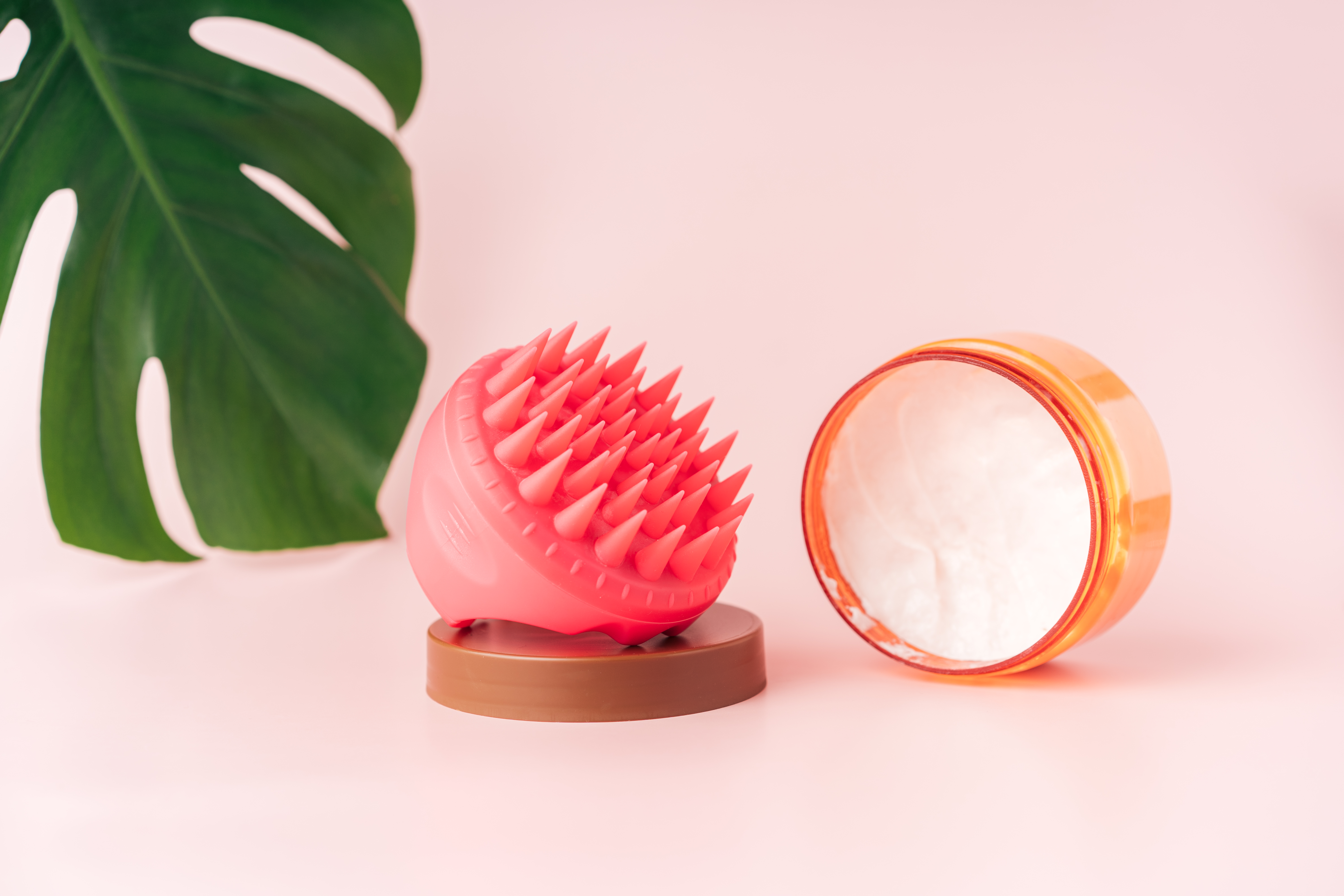 Head massager and head cream against pink background and monstera leaf Head massager and head cream against pink background and monstera leaf