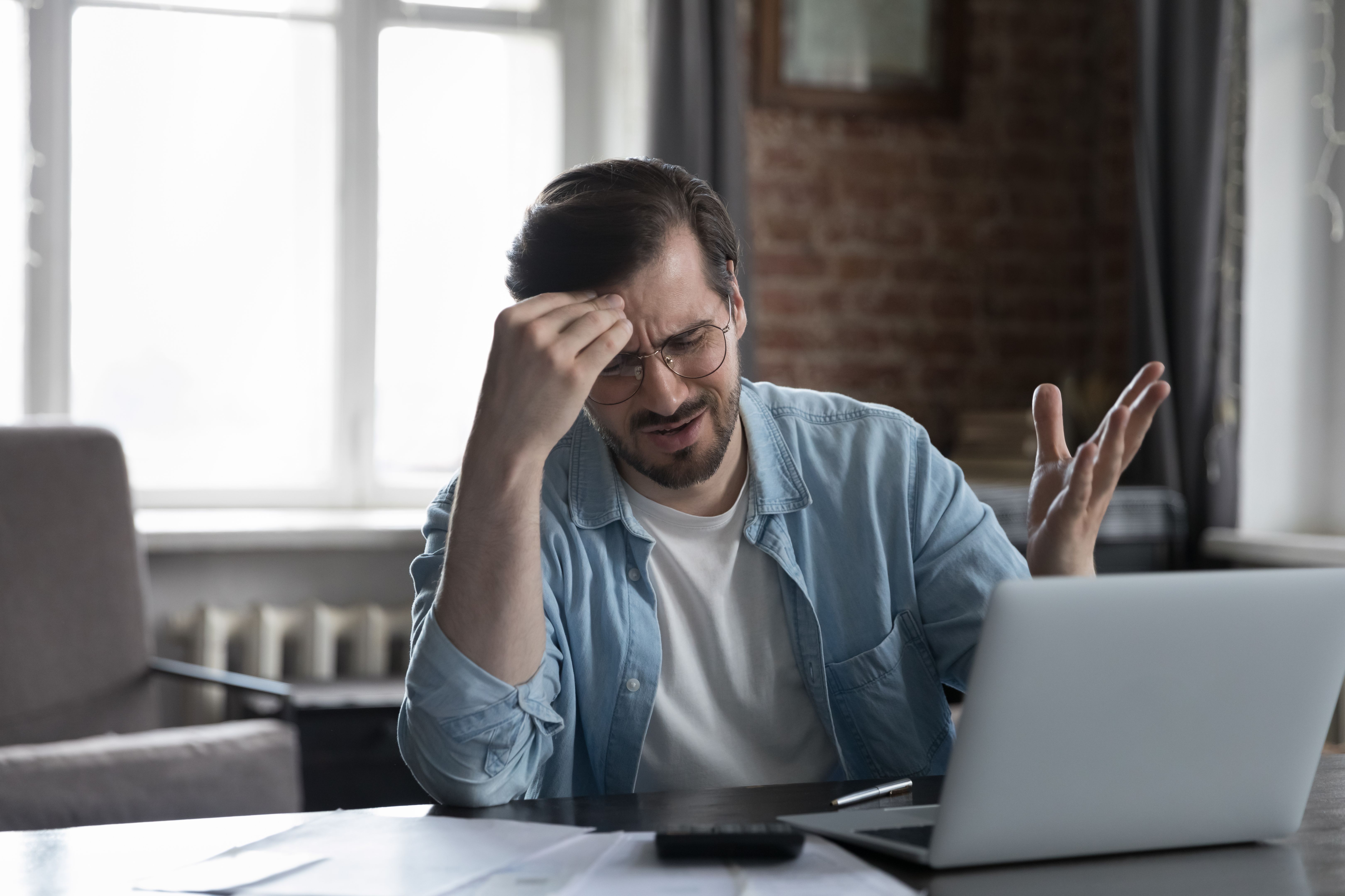 Desperate upset laptop user man sitting at paper bills, documents
