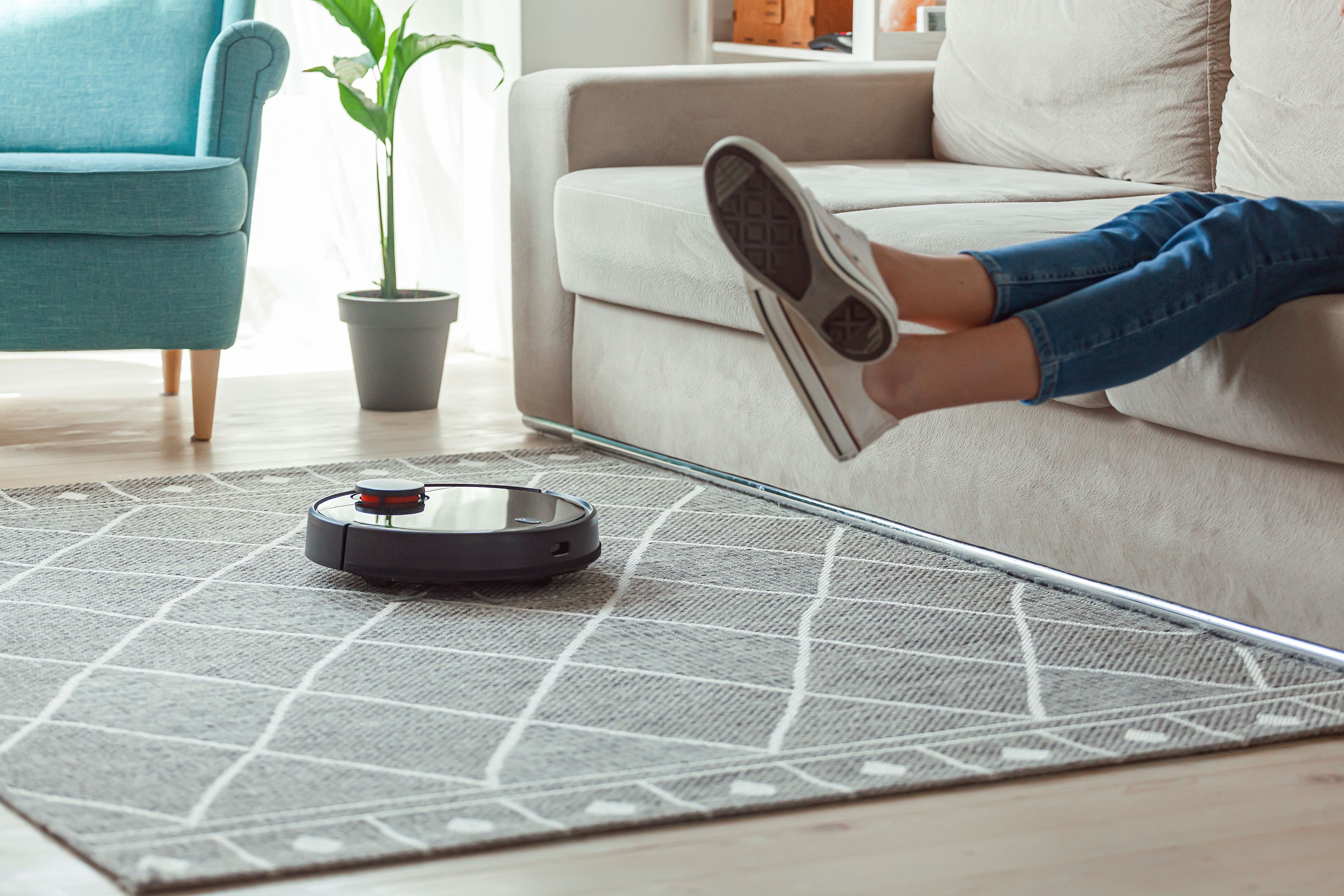Robot vacuum cleaner cleaning carpet, woman legs rest sitting on sofa at home