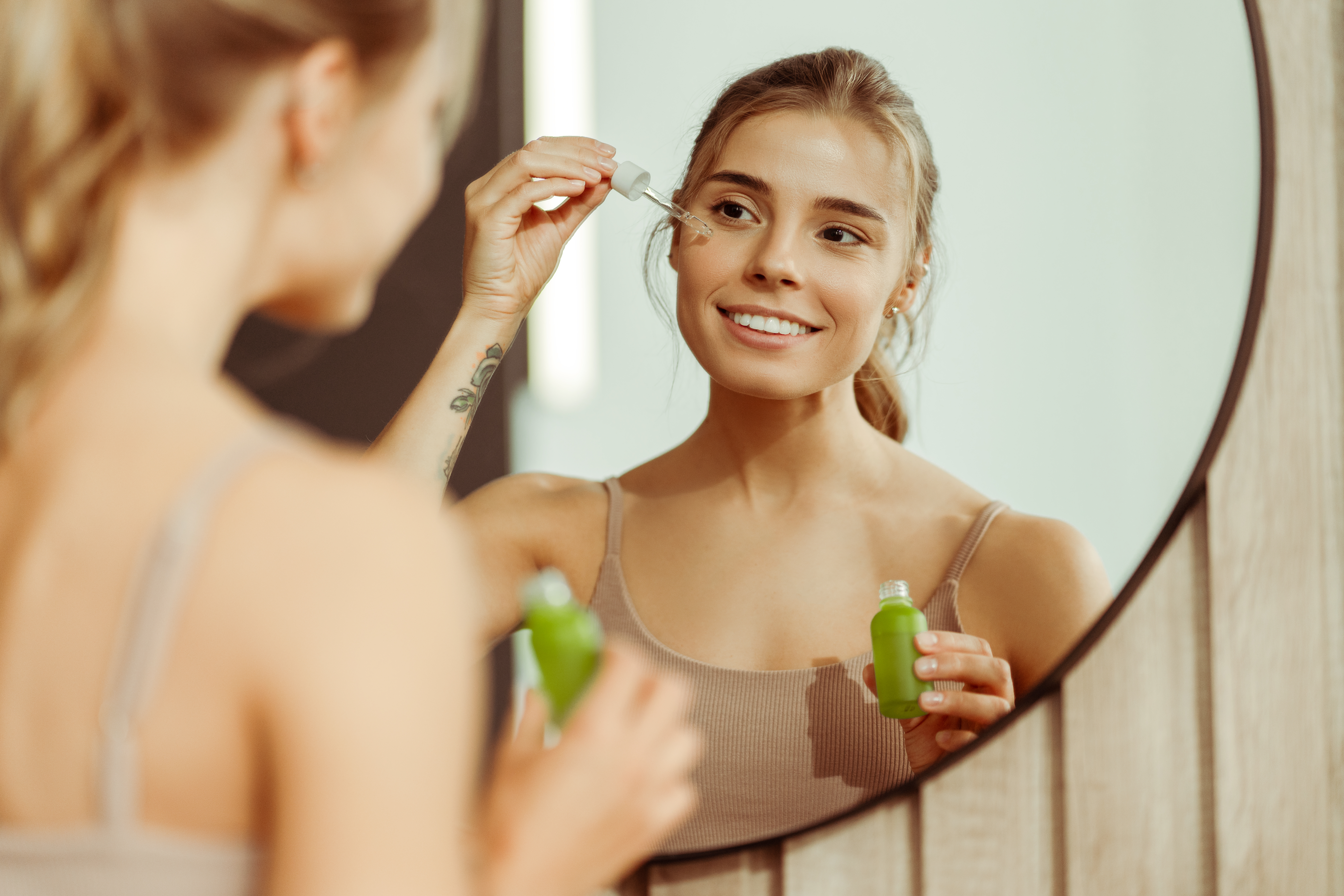 Young woman holding green bottle applying serum to face, looking in mirror, standing in bathroom Young woman holding green bottle applying serum to face, looking in mirror, standing in bathroom