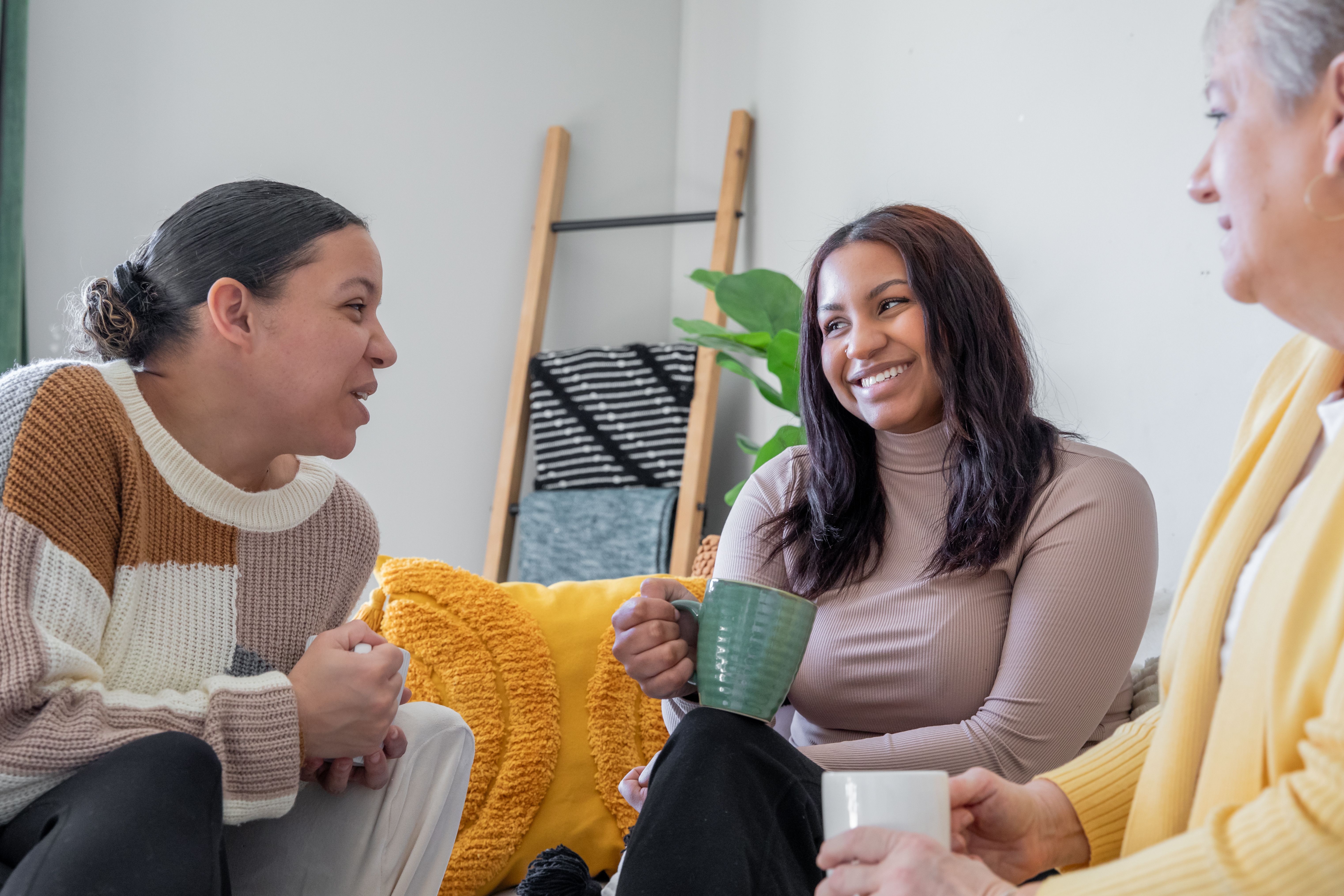 Three women, seated on a couch, enjoy a lively conversation over mugs of coffee.