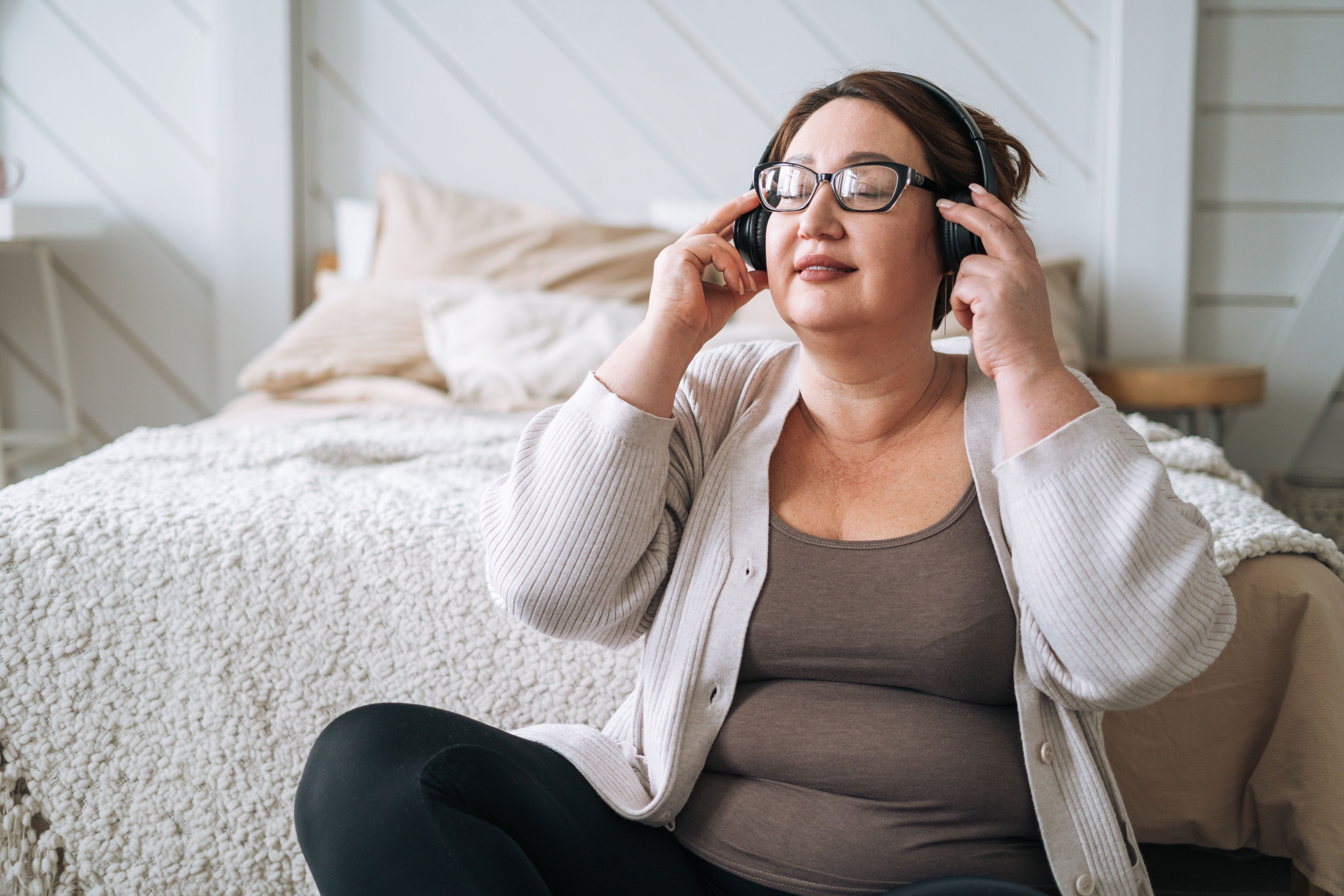 Middle aged plus size smiling brunette woman in home wear listening music in headphones in bedroom