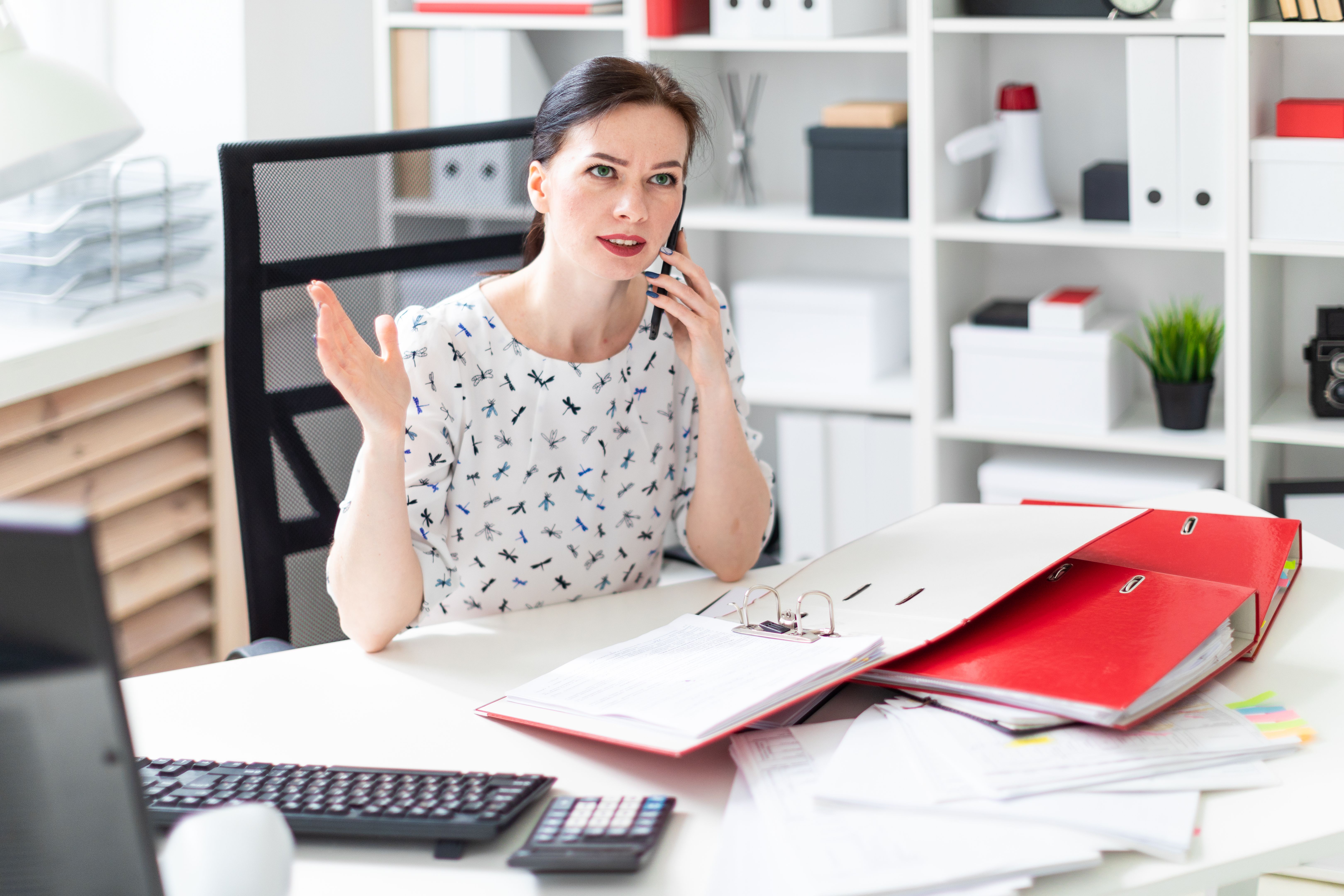 A young girl sitting in the office at the computer Desk, working with documents and talking on the phone.
