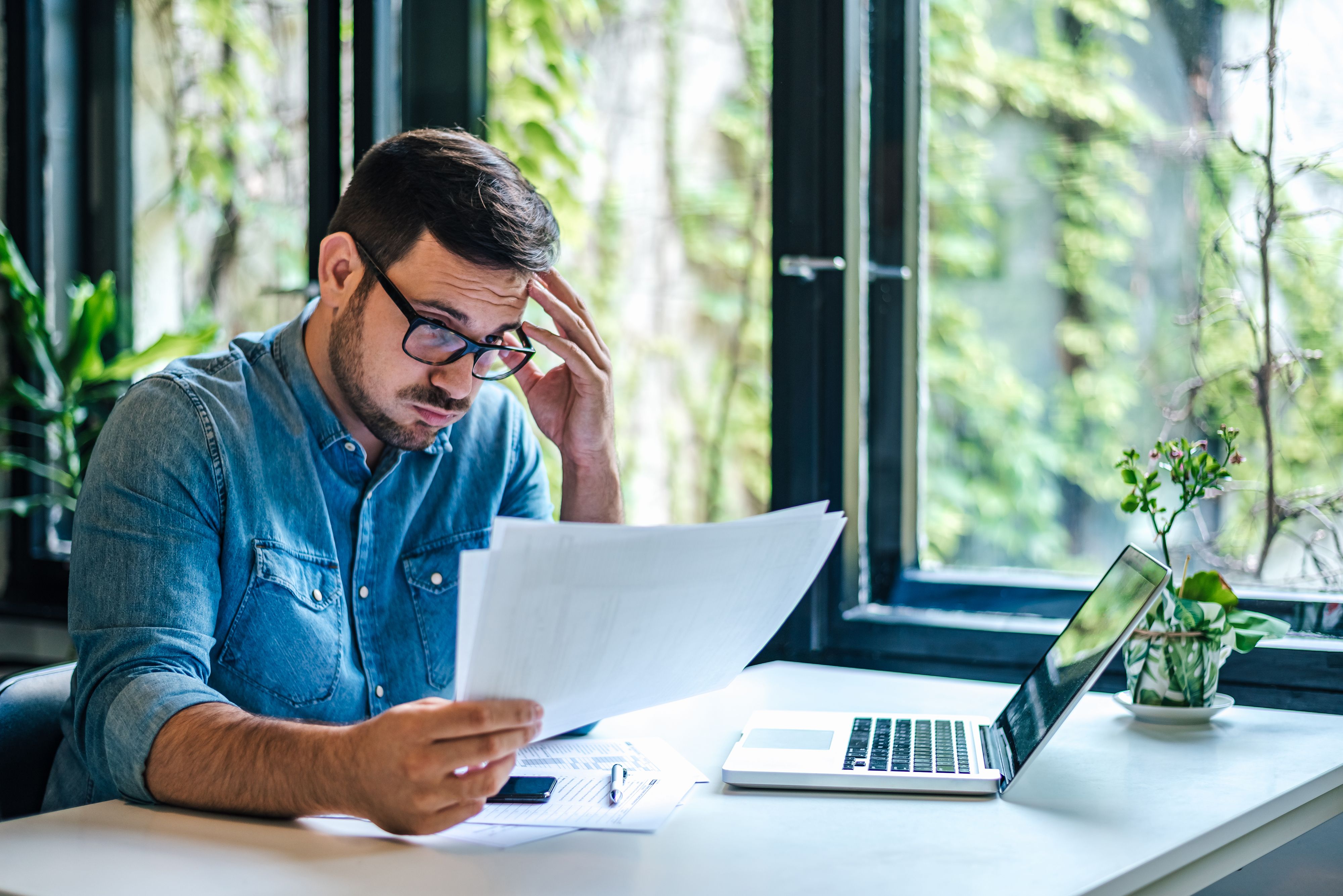 Stressed businessman reading documents while working on laptop at home office Stressed businessman reading documents while working on laptop at home office