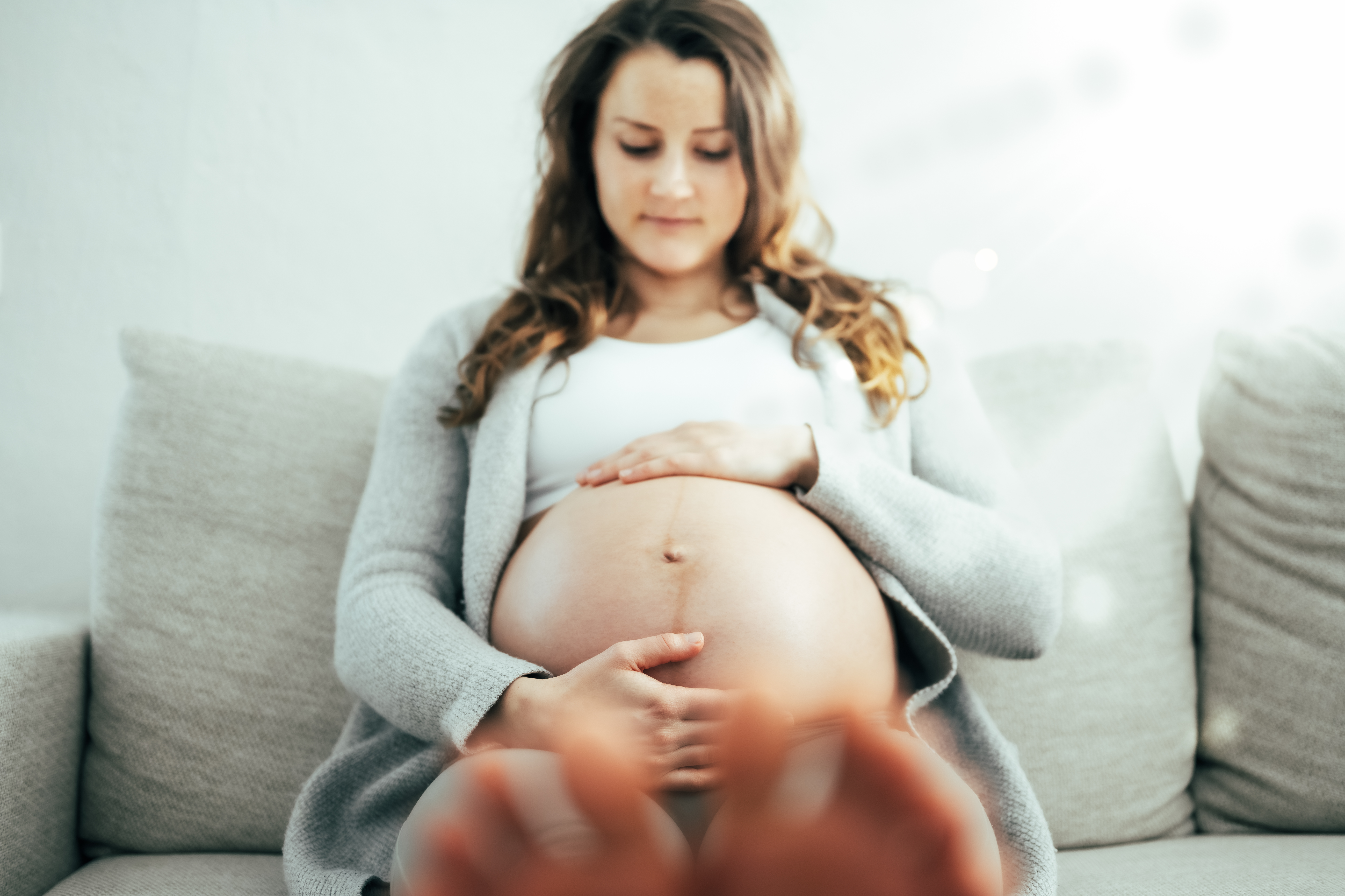Frontal view of happy woman sitting on sofa gently holding her belly in imagination of baby in last stage of pregnancy. Pregnancy third trimester - week 34.