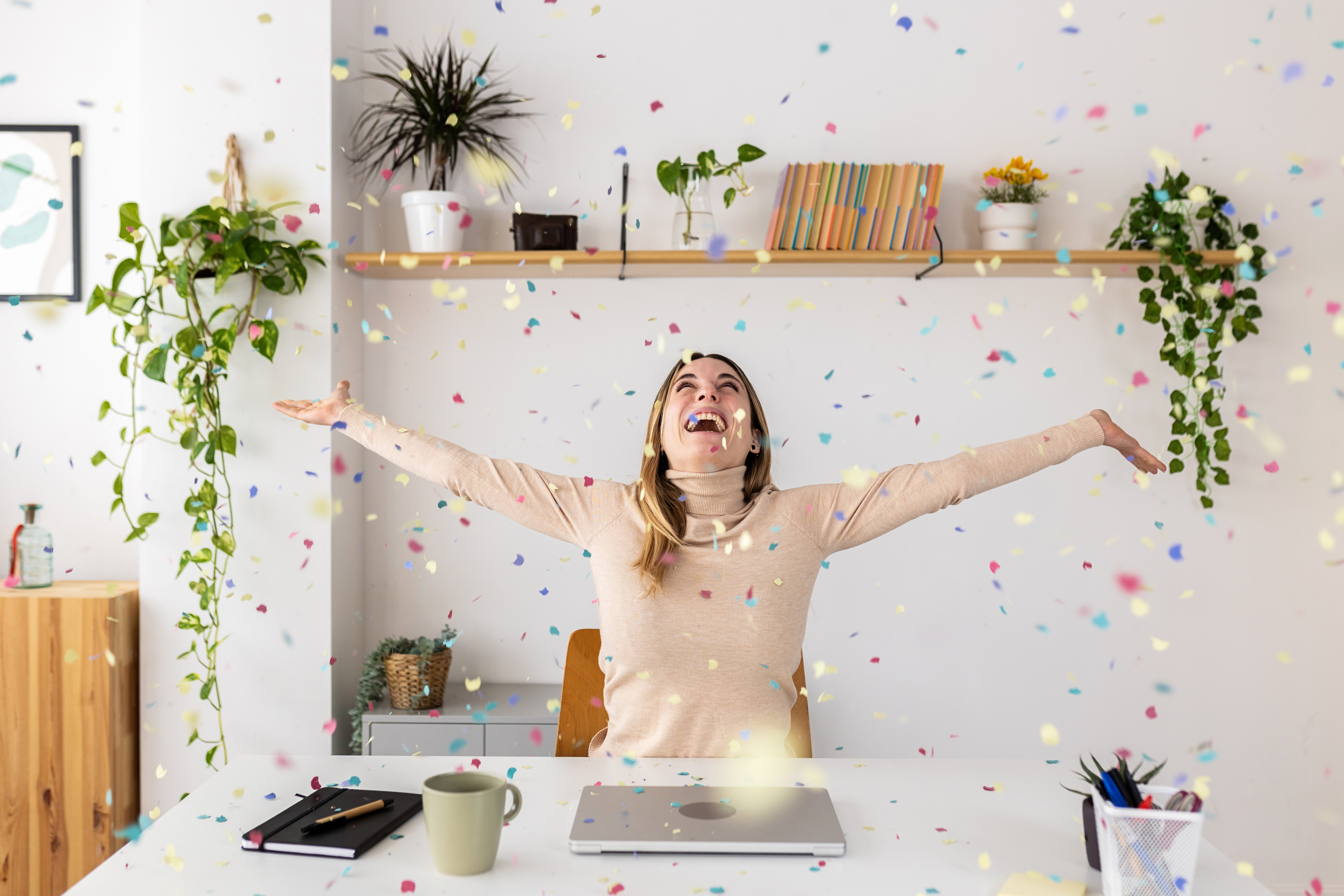 Young businesswoman celebrating success throwing confetti sitting at home office