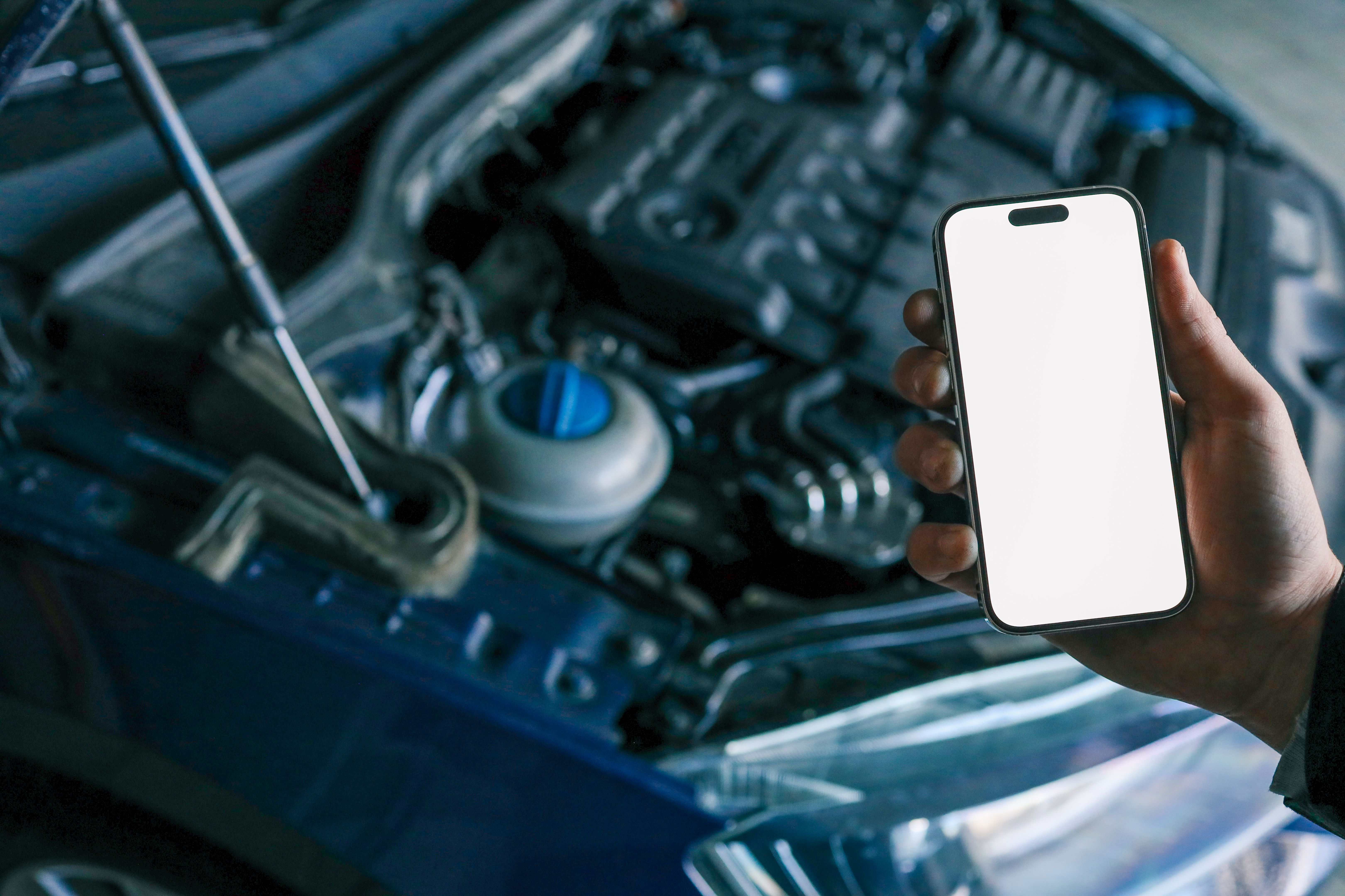 Hand holding a smartphone with a blank screen in front of an open car hood, showing the engine compartment. Concept for car diagnostics, automotive apps, or remote vehicle monitoring Hand holding a smartphone with a blank screen in front of an open car hood, showing the engine compartment. Concept for car diagnostics, automotive apps, or remote vehicle monitoring