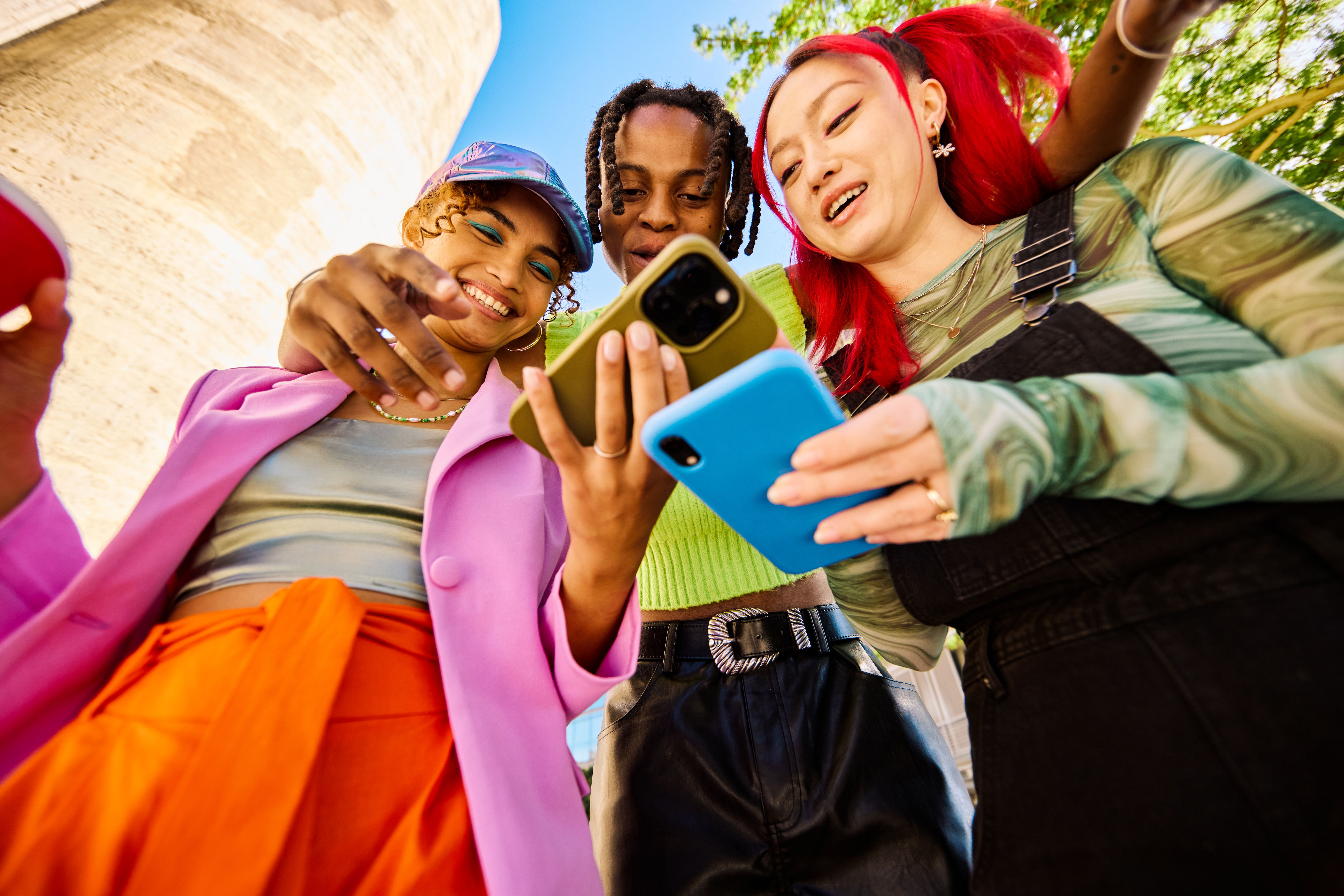 Three Gen Z adults smiling and looking at smartphones outdoors in an urban environment. Low angle shot. Social media and mobile finance themes.