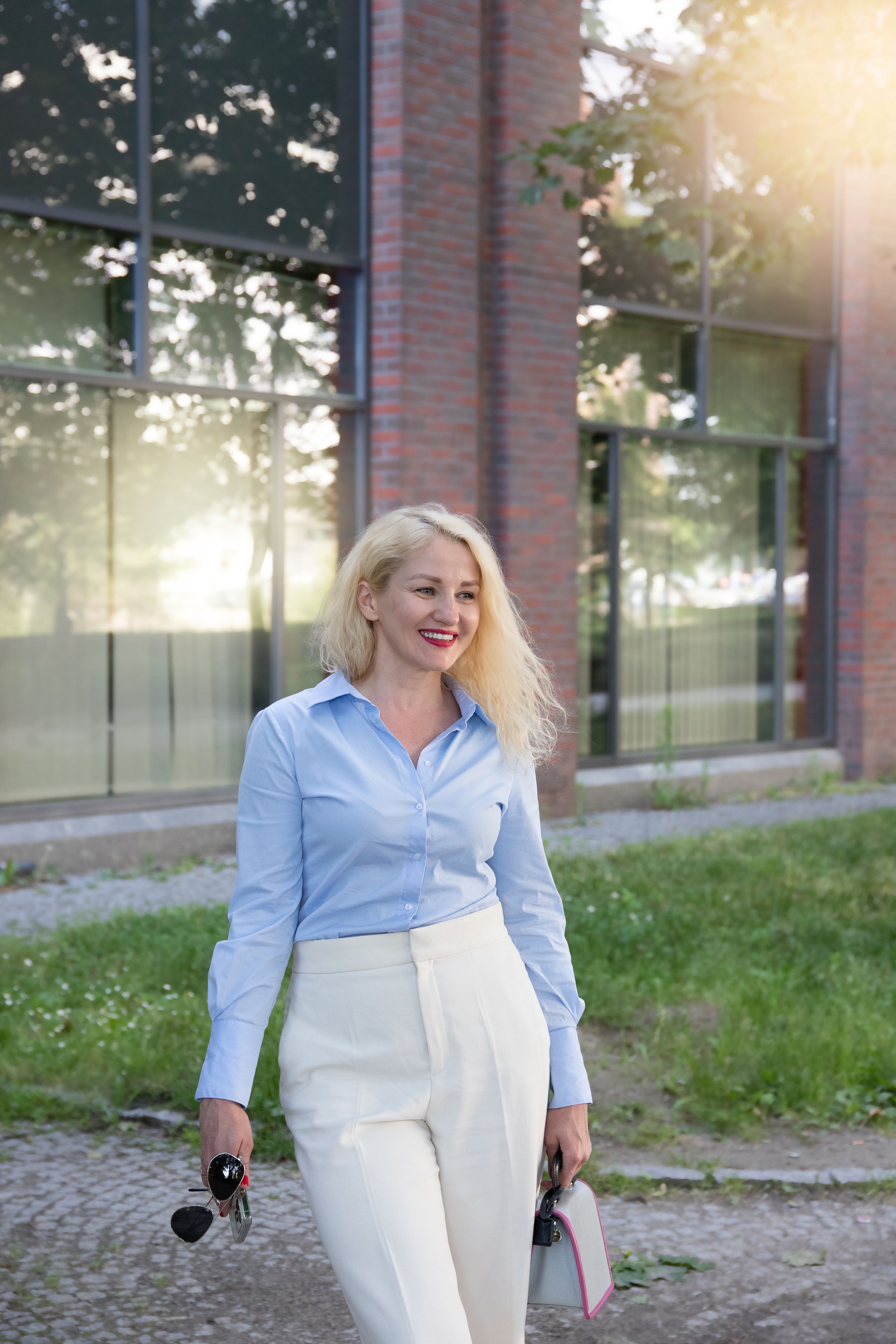 A successful businesswoman in a blue shirt and white trousers is standing outside the office. A warm sunny day. A successful businesswoman in a blue shirt and white trousers is standing outside the office. A warm sunny day.