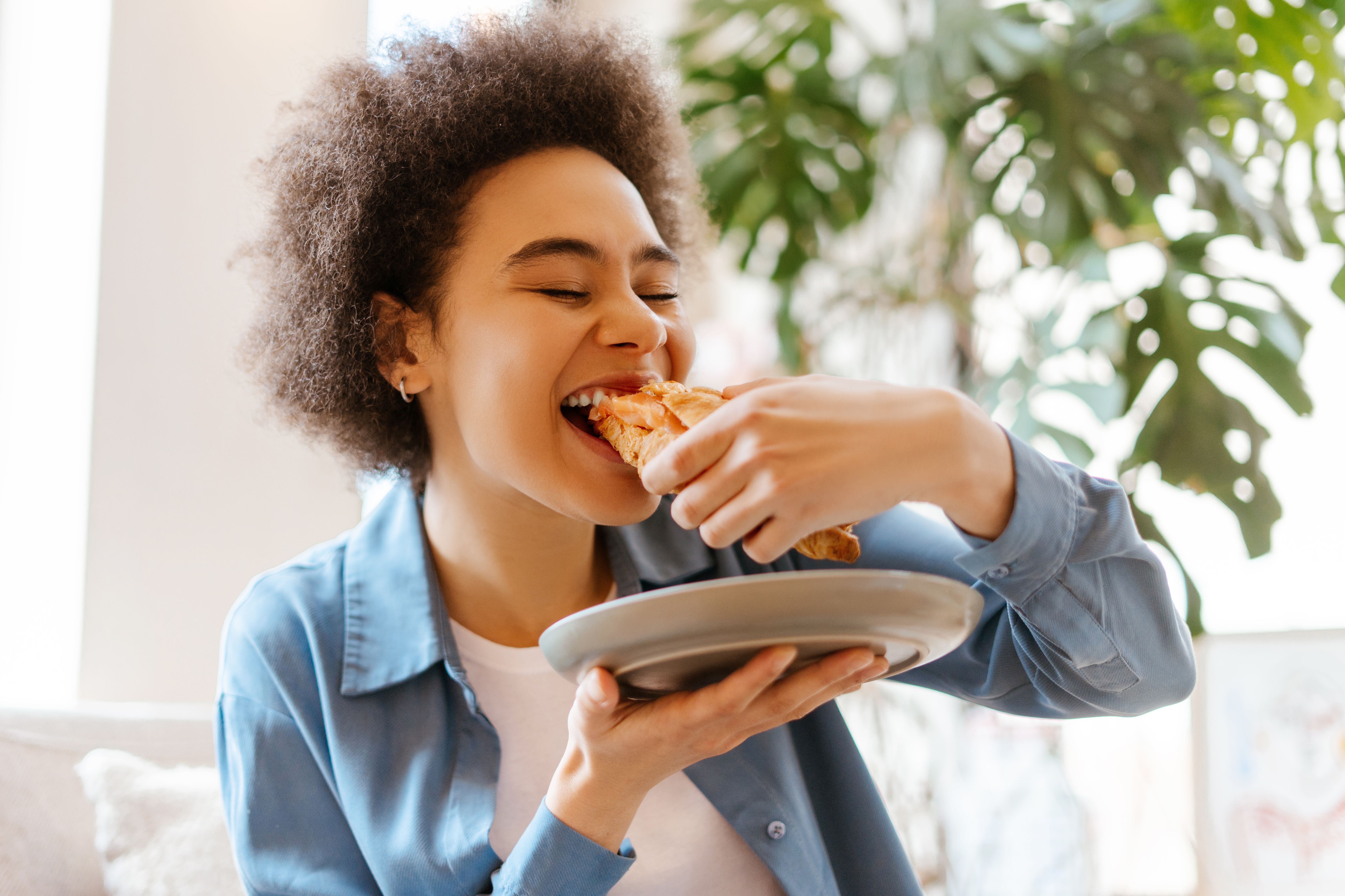 Beautiful, authentic African American young woman eating fresh croissant, eyes closed, enjoying Beautiful, authentic African American young woman eating fresh croissant, eyes closed, enjoying